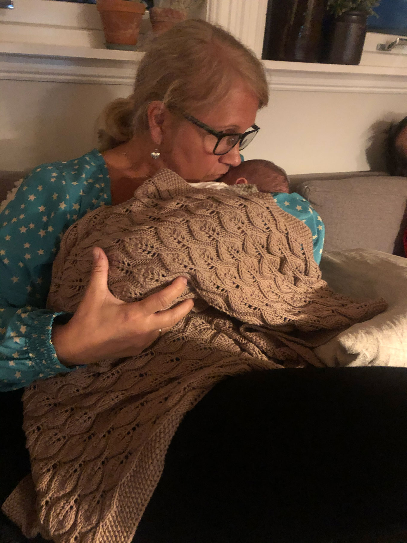 Kvinne i slutten av 50-årene med briller holder en baby pakket inn i et strikketeppe. Hun har briller på seg og beundrer den lille.