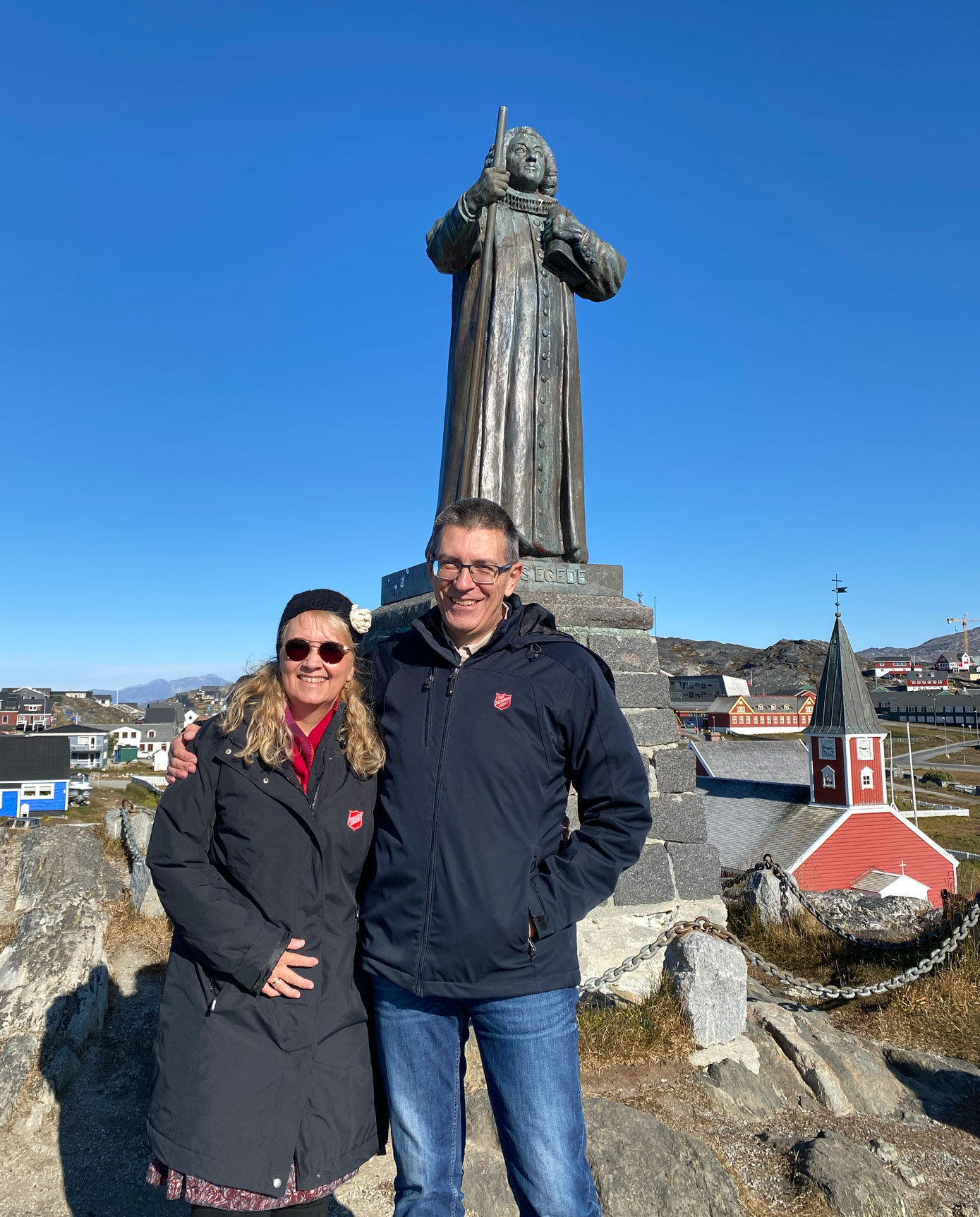 Kvinne og mann står foran en statue. Himmelen er knall blå og det er fargerike hus i bakgrunnen, i rødt og blått.