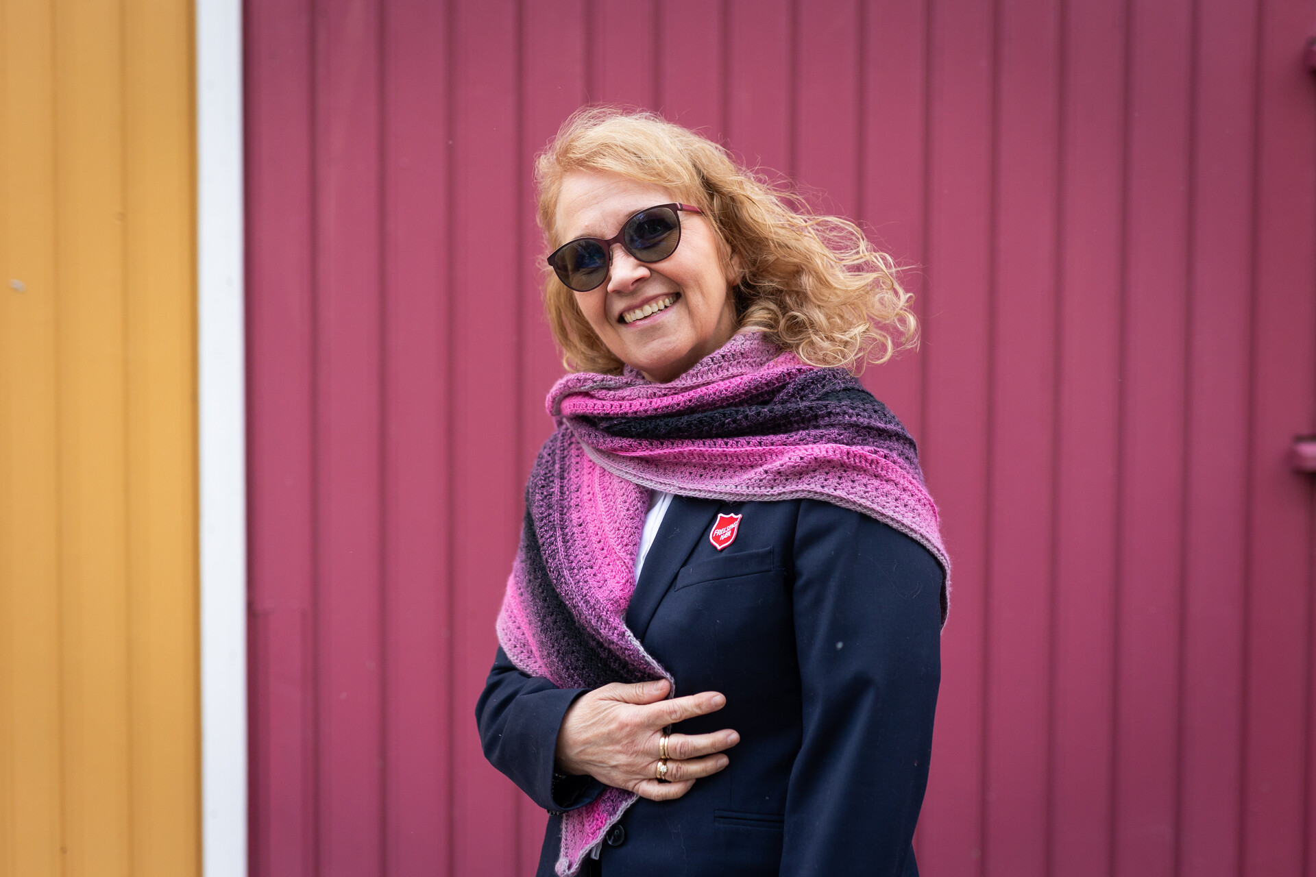 Foto: Kvinne tidlig i 60-årene med ullskjerf i rosa og lilla står mot en rosa og gul husvegg.