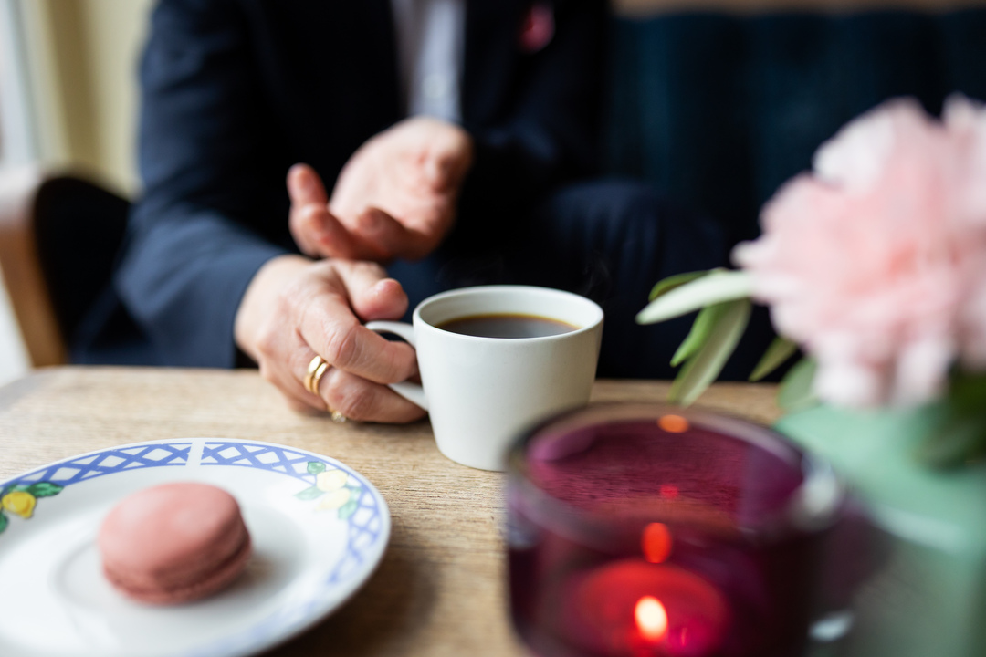 Foto: En kvinnes hender holder en kopp med svart kaffe. VI ser ikke ansiktet. På bordet står blomster, et lys og det ligger en rosa makron på bordet.