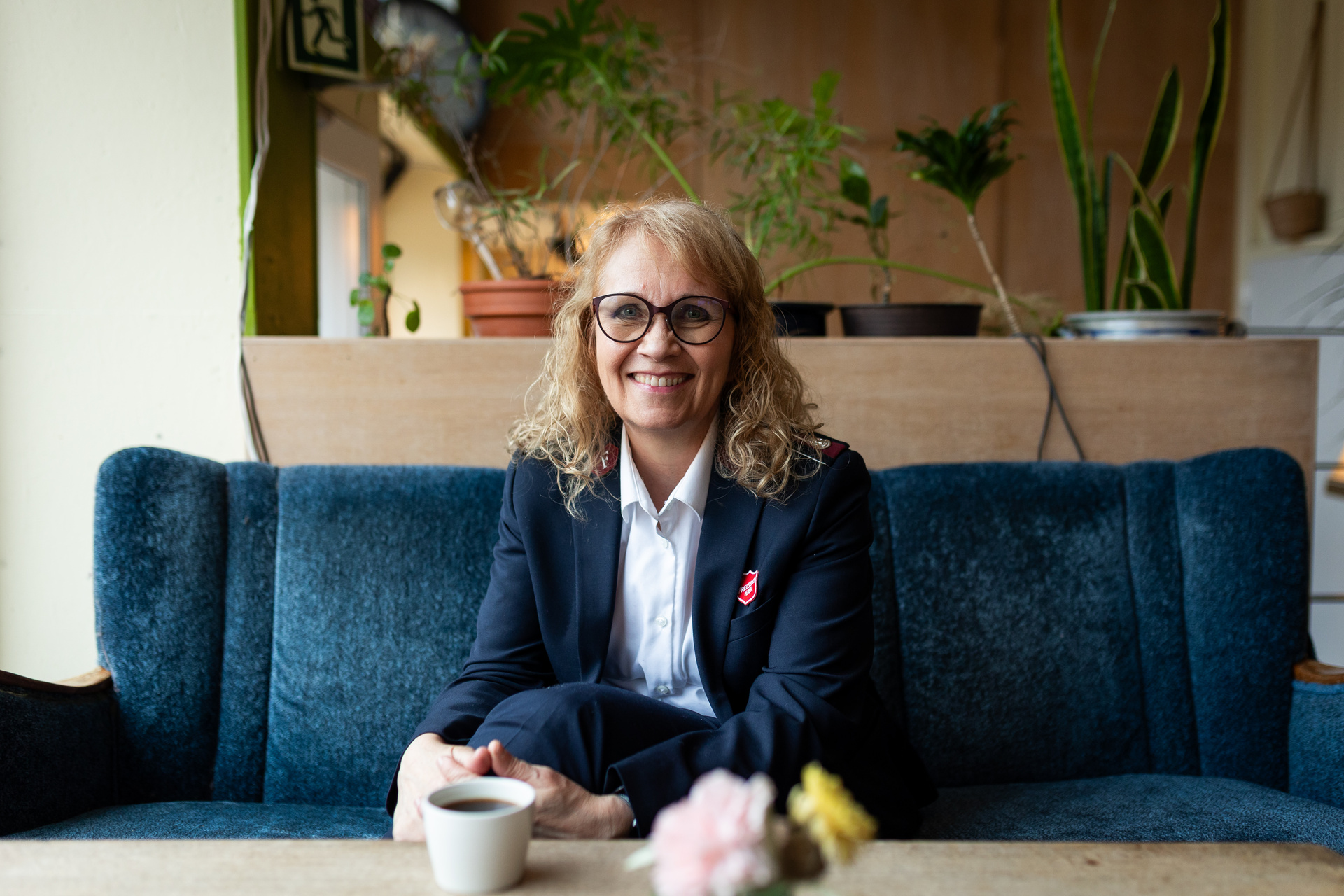 Kvinne med briller, krøller og uniform fra Frelsesarmeen sitter i en blå sofa. På bordet foran hennes står det en kopp med kaffe og noen blomster. I bakgrunnen er det grønne planter og hyller på en vegg bak.
