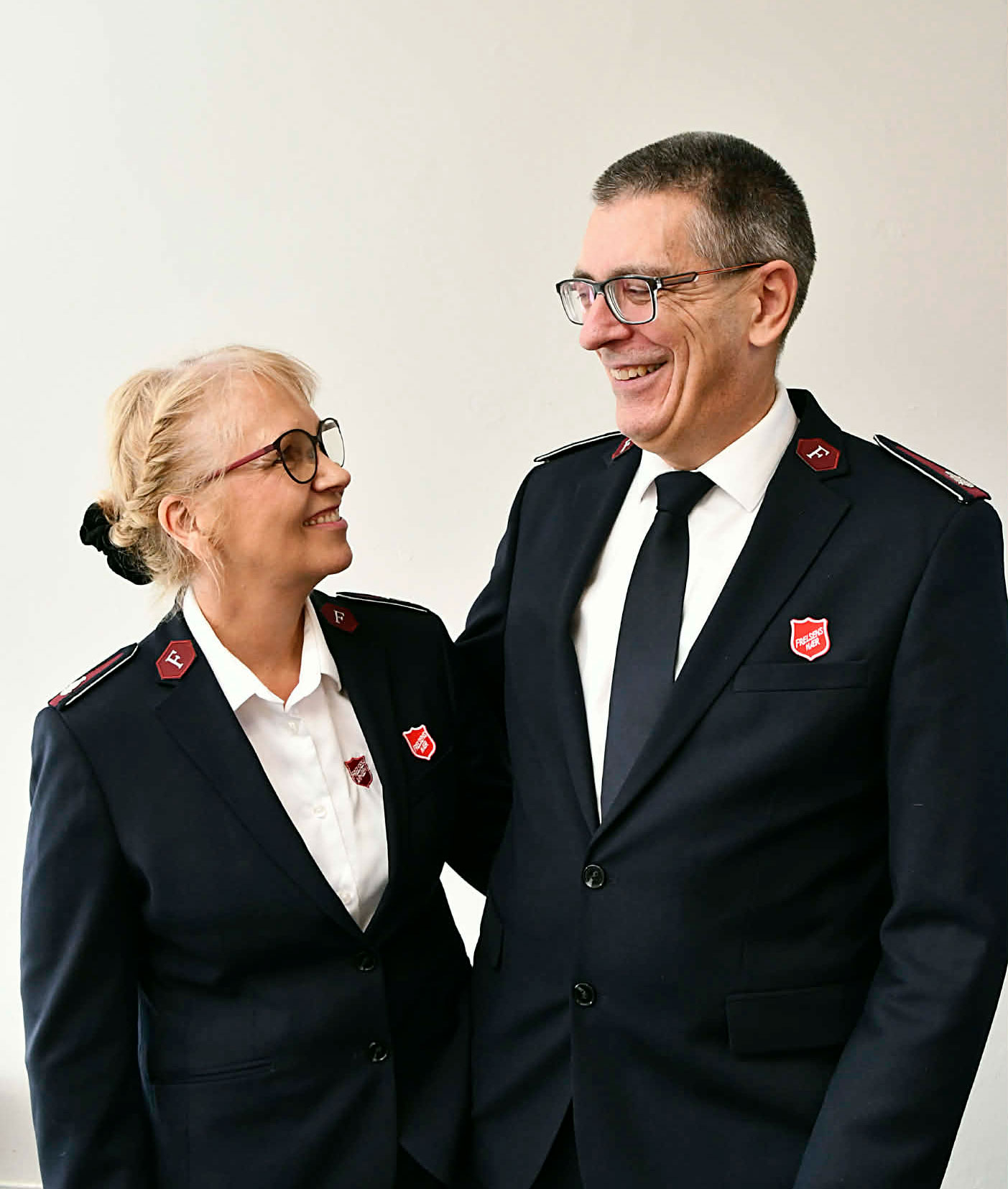Kvinne og mann i uniformer fra Frelsesarmeen ser på hverandre. De er tidlig i 60-årene og han er mye høyere enn henne.