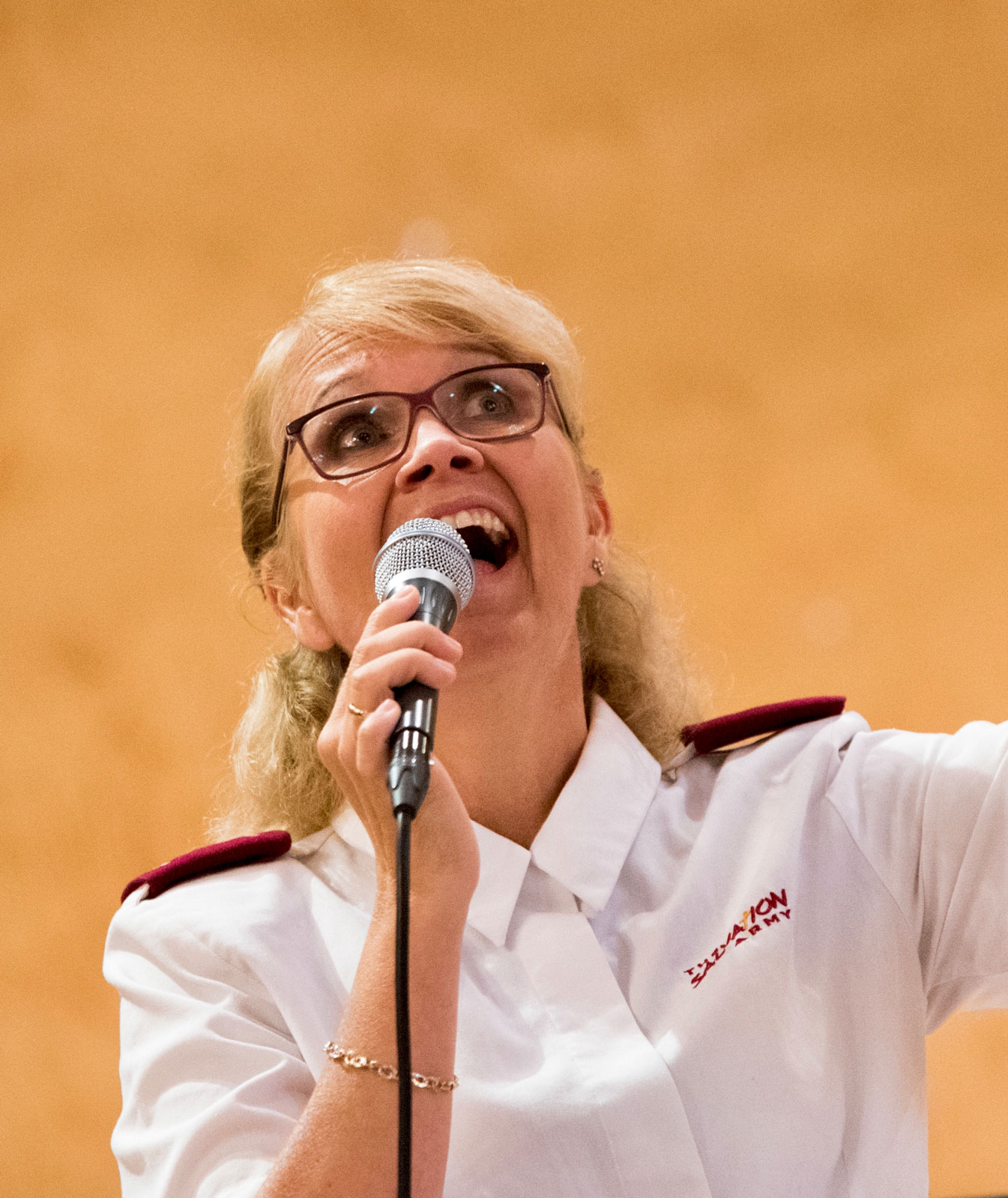 Blond kvinne i 40-årene i hvit uniformsskjorte fra Frelsesarmeen syngeri en mikreofon og har blikket rettet oppover. Veggen bak er gul og dette er minst 20 år tilbake i tid.