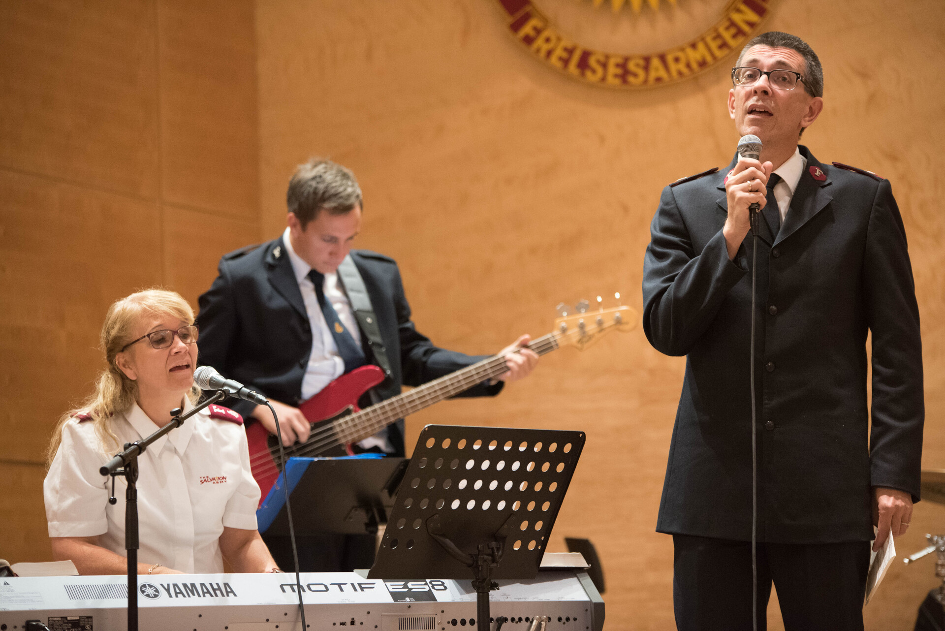 Foto: I et menighetsrom sitter en kvinne i 50-årene bak et keyboard og synger. Bak spiller en mann i midten av 20-årene rød gitar og foran igjen står en mann i 50-årene med en mikrofon og synger.  Alle har på seg uniformer fra Frelsesarmeen og de to eldste har briller.