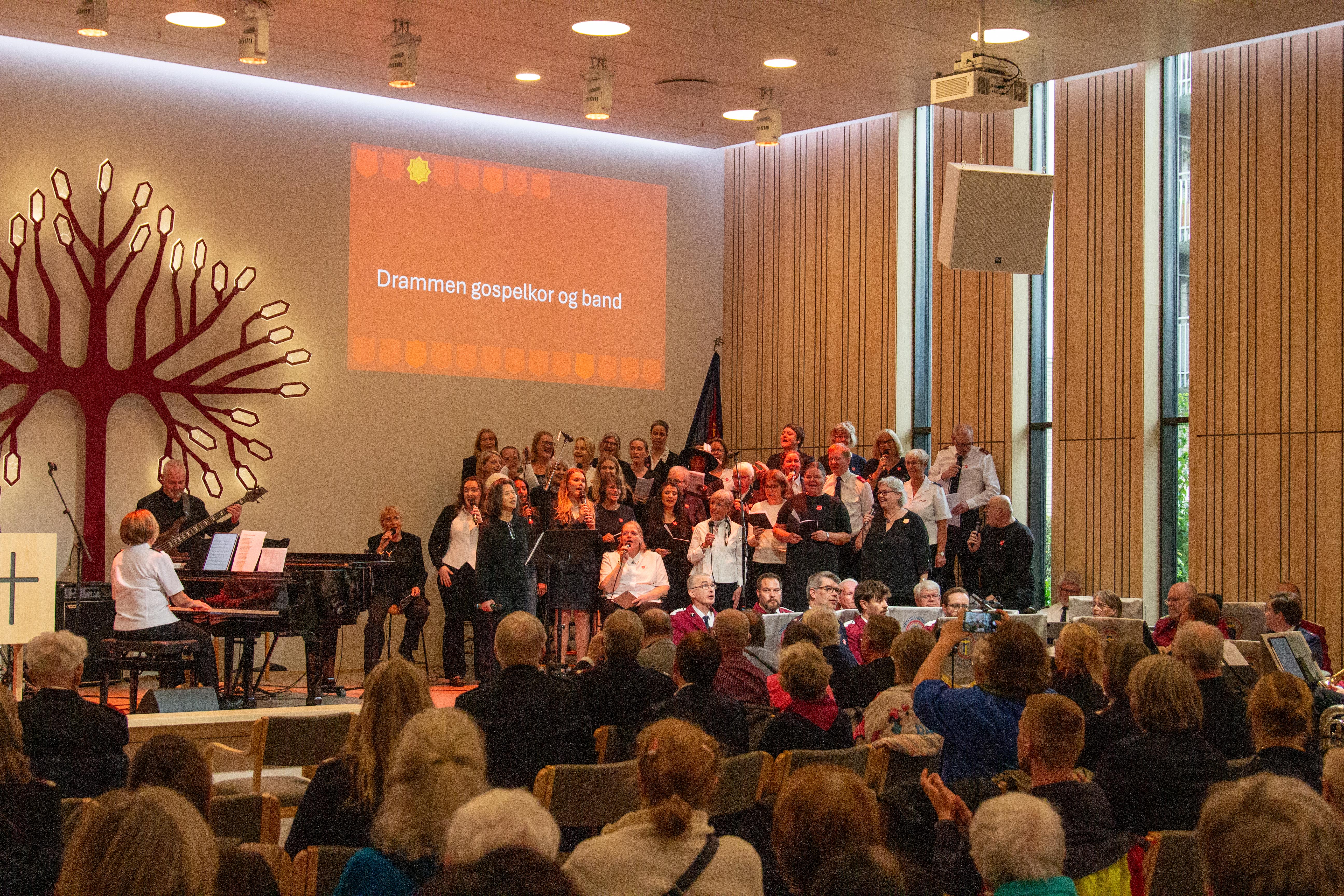Drammen gospelkor synger på posiet ved Frelsesarmeens korps på Ensjø. Beate Holm Hansen sitter ved flygelet, og vi ser frelsesarmeens territoriale hornorkester foran koret samt publikum i forgrunnen av bildet.