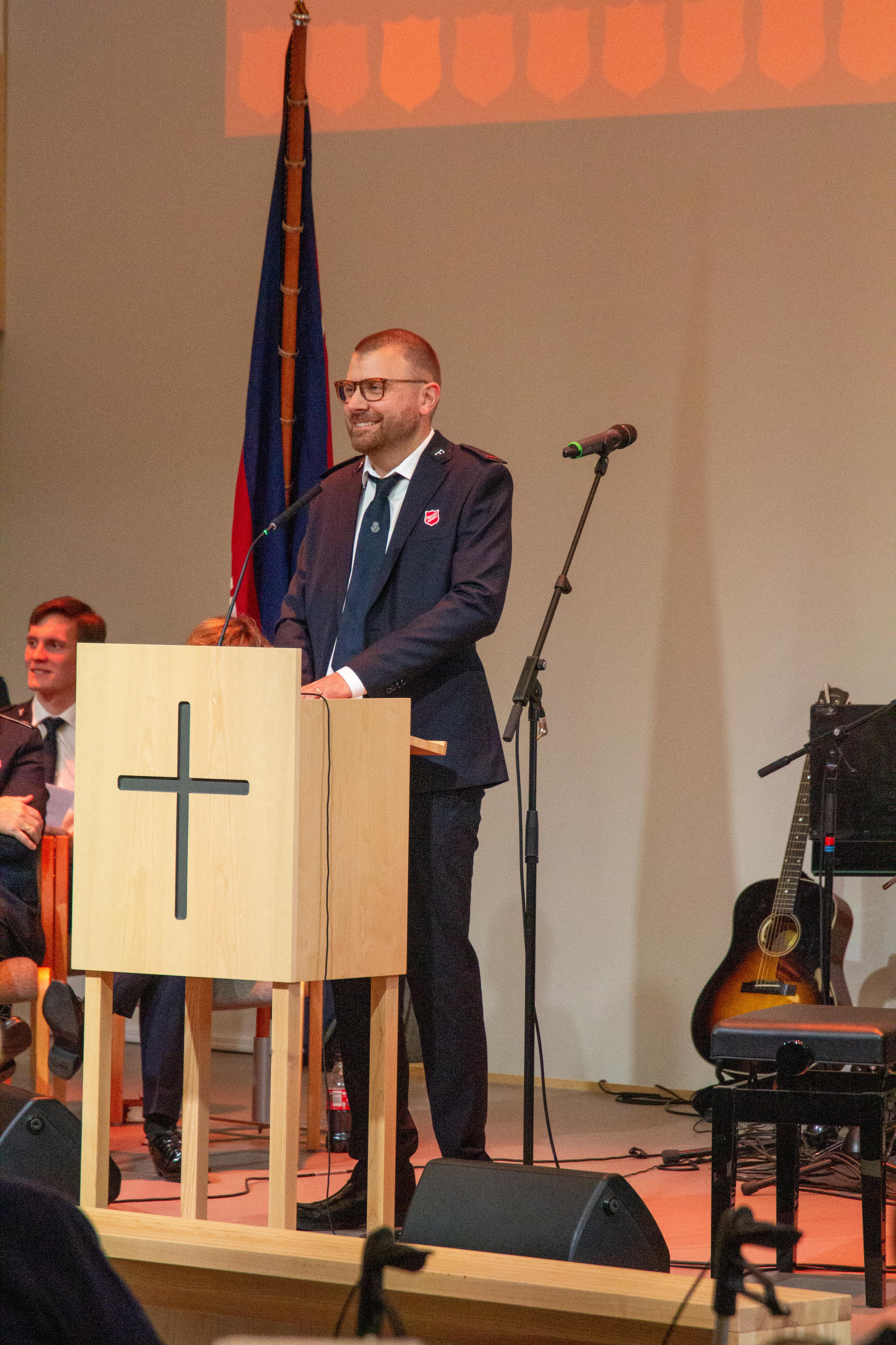 Kadett Sam Ruck-Penderstår på scenen i Ensjø korps bak en trehvit talerstol med sølvkors på. Bak ham ser vi Frelsesarmeens flagg.