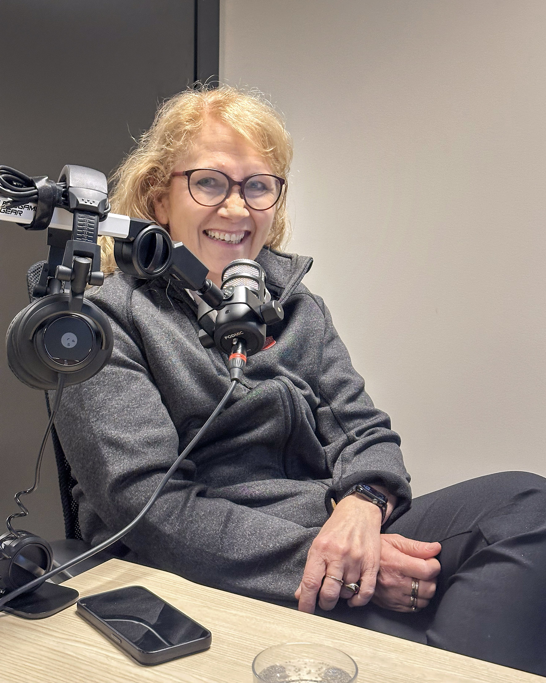 Foto: Blond kvinne i 60-årene sitter og ler foran en mikrofon i et lydstudio. Hun har på seg grå flisjakke med logo fra Frelsesarmeen.
