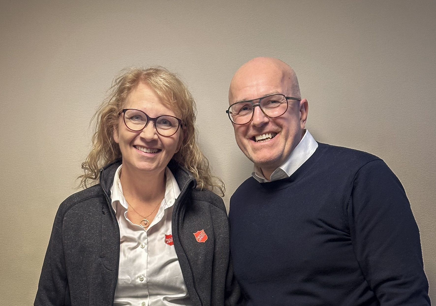 Foto: Kvinne i 60-årene og mann i 50-årene ler inn i kamera. Begge har briller og klær fra Frelsesarmeen. Hun er blond og han har glattbarbert hode.