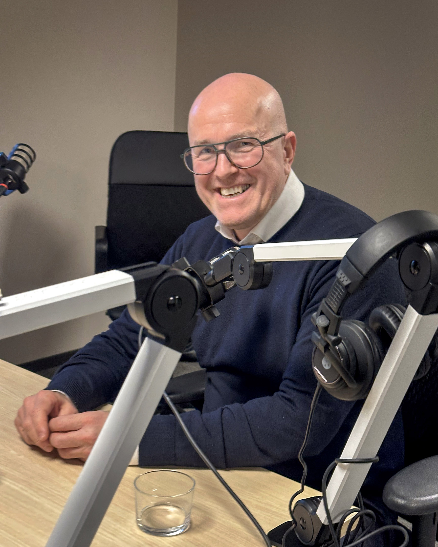 Foto: Mann i 50-årene med glattbarbert hode og briller sitter foran en mikrofon i et lydstudio og smiler bredt slik at alle tennene viser. Han har på seg en mørkeblå genser over en hvit skjorte.