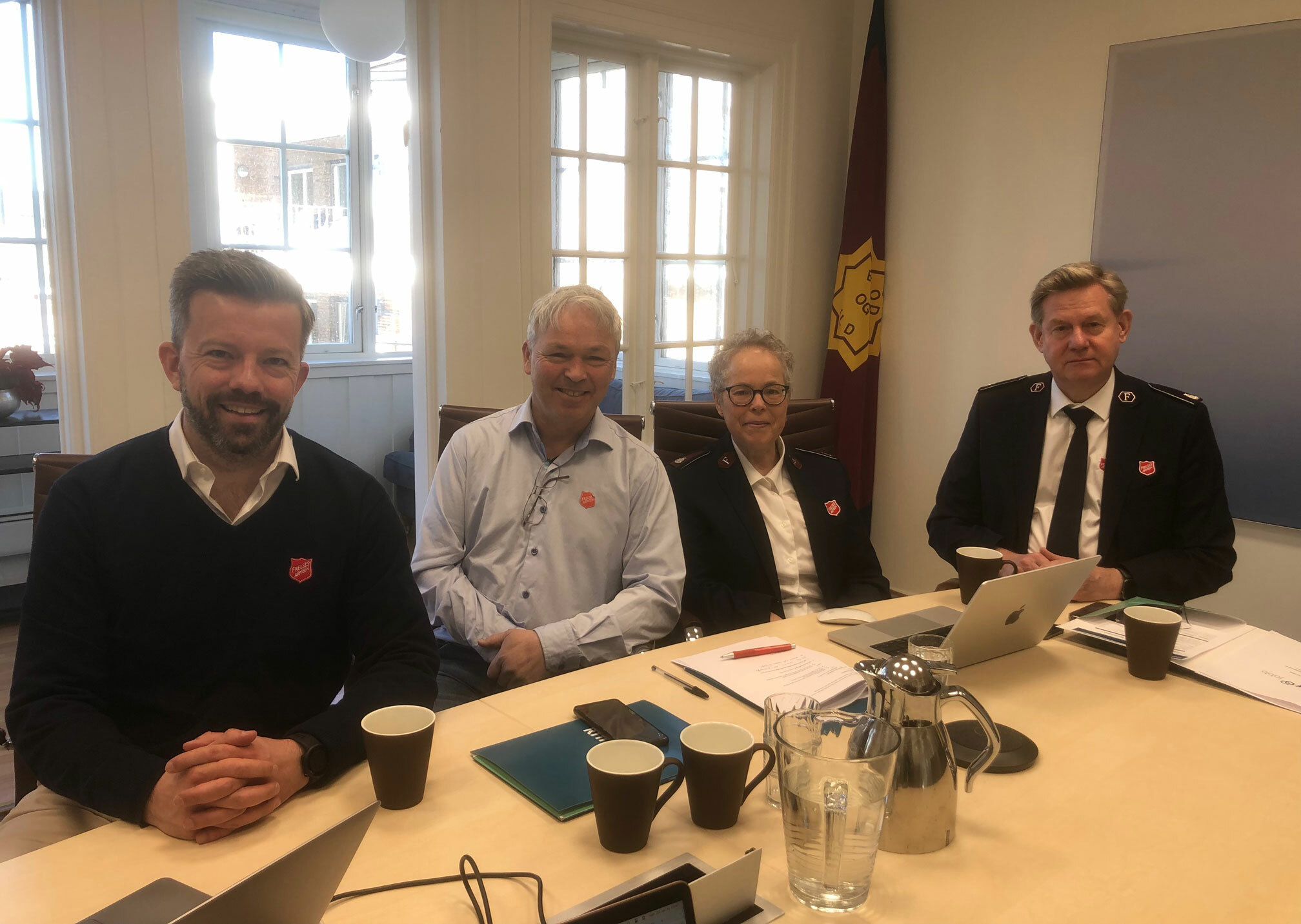 Foto: Tre menn og en kvinne sitter på rekke ved et bord. To av dem har på seg uniform fra Frelsesarmeen.