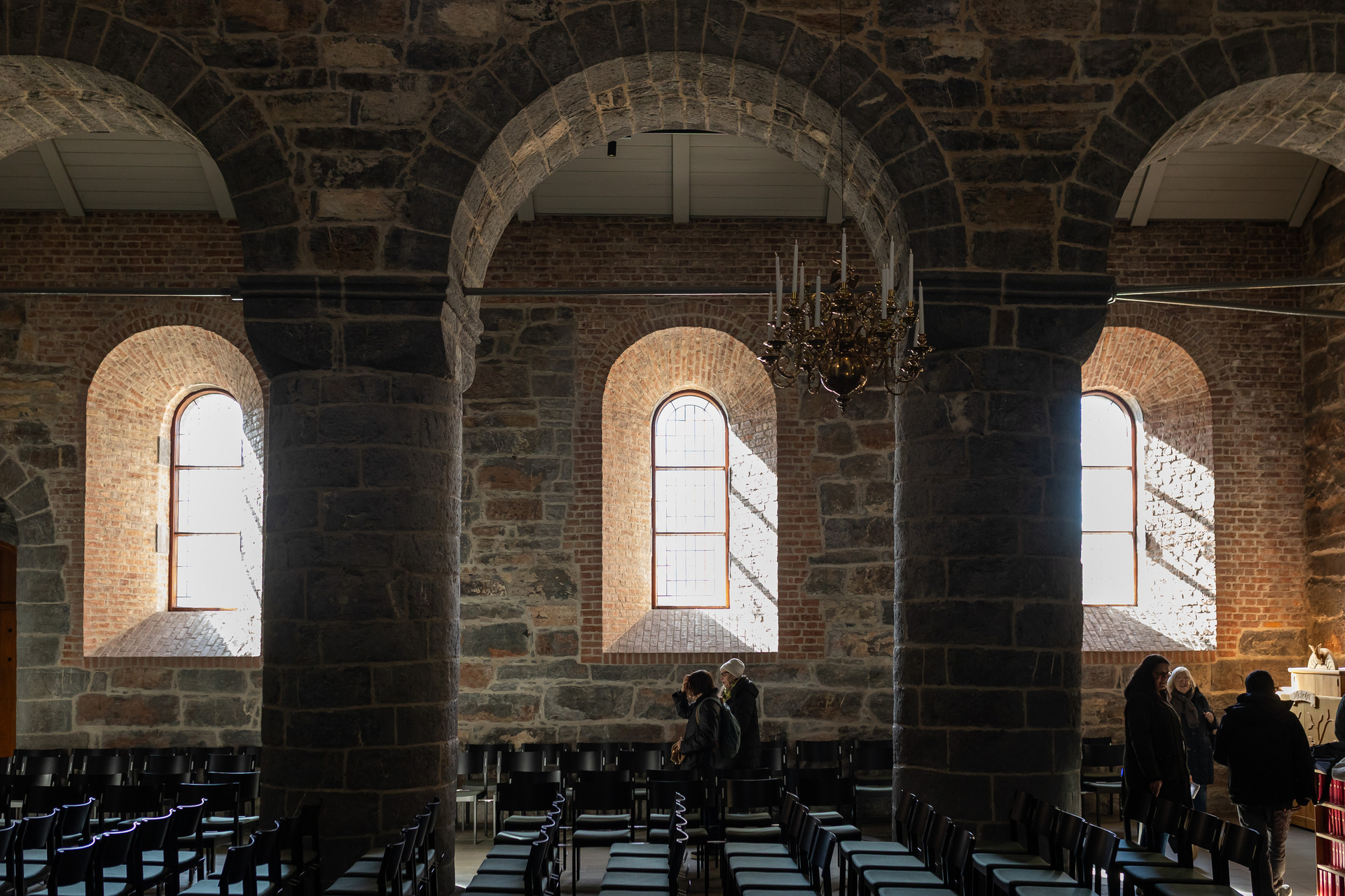 Foto: Bildet er tatt inne i en gammel kirke. I forgrunnen sees tre bueganger med søyler. Bag dem er tre buede vinduer som slipper dagslys inn i et ganske mørkt rom. Noen mennesker skimtes i bakgrunnen,