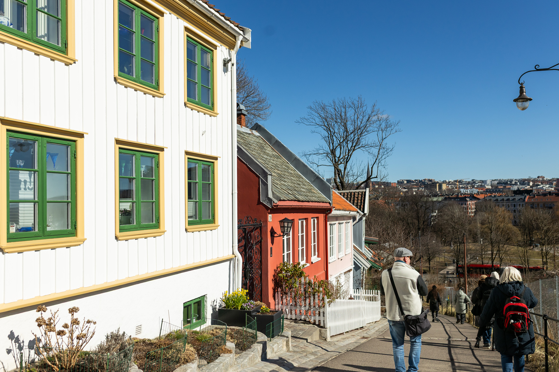 Foto: En gruppe mennesker sees bakfra på litt avstand. De er på vei ned en gate med gamle trehus på den ene siden. Over dem er en blå og skyfrie himmel.