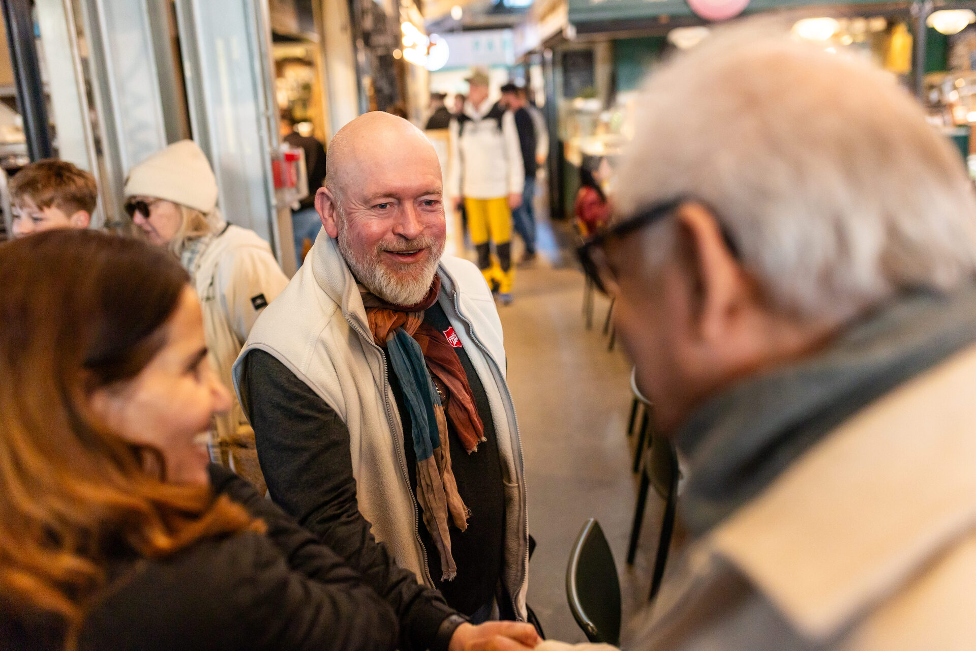 Foto: En skallet mann med grått skjegg lener seg litt fram og håndhilser på en annen mann. Den andre mannen sees uskarpt med ryggen til han har grått hår og briller. En kvinne med brunt hår står også me ryggen til, hun vender seg mot mannen og smiler.