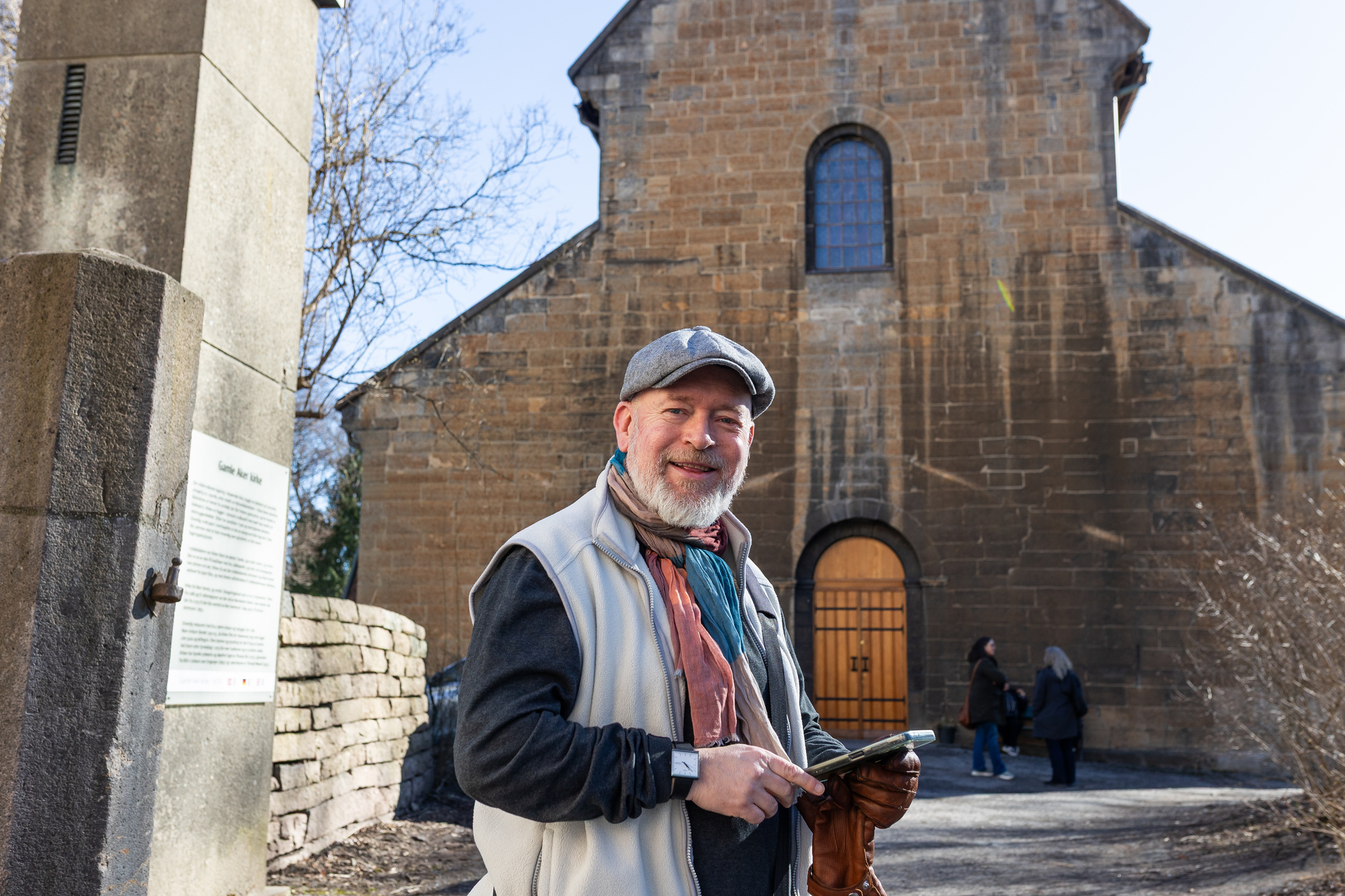 Foto: En mann med grått skjegg, sixpense og grå vest over en svart genser står og smiler mot kamera. Bak ham sees deler av en gammel murkirke, med inngangsdør i tre.