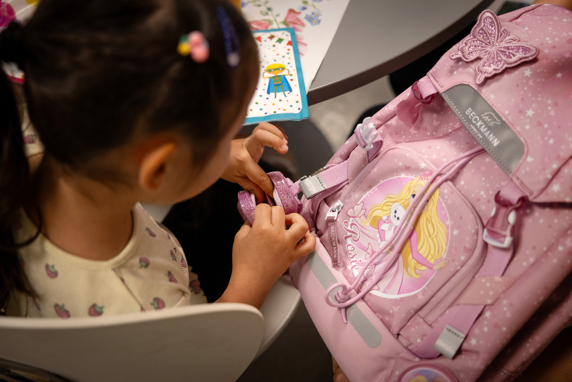 Foto: Ei jente med mørkt langt hår sees uskarpt bakfra. Foran henne er en rosa sekk med en prinsesse og sommerfuglpynt på. Huin holder på å åpne en liten glitrende sekk som henger på siden av skolesekken.