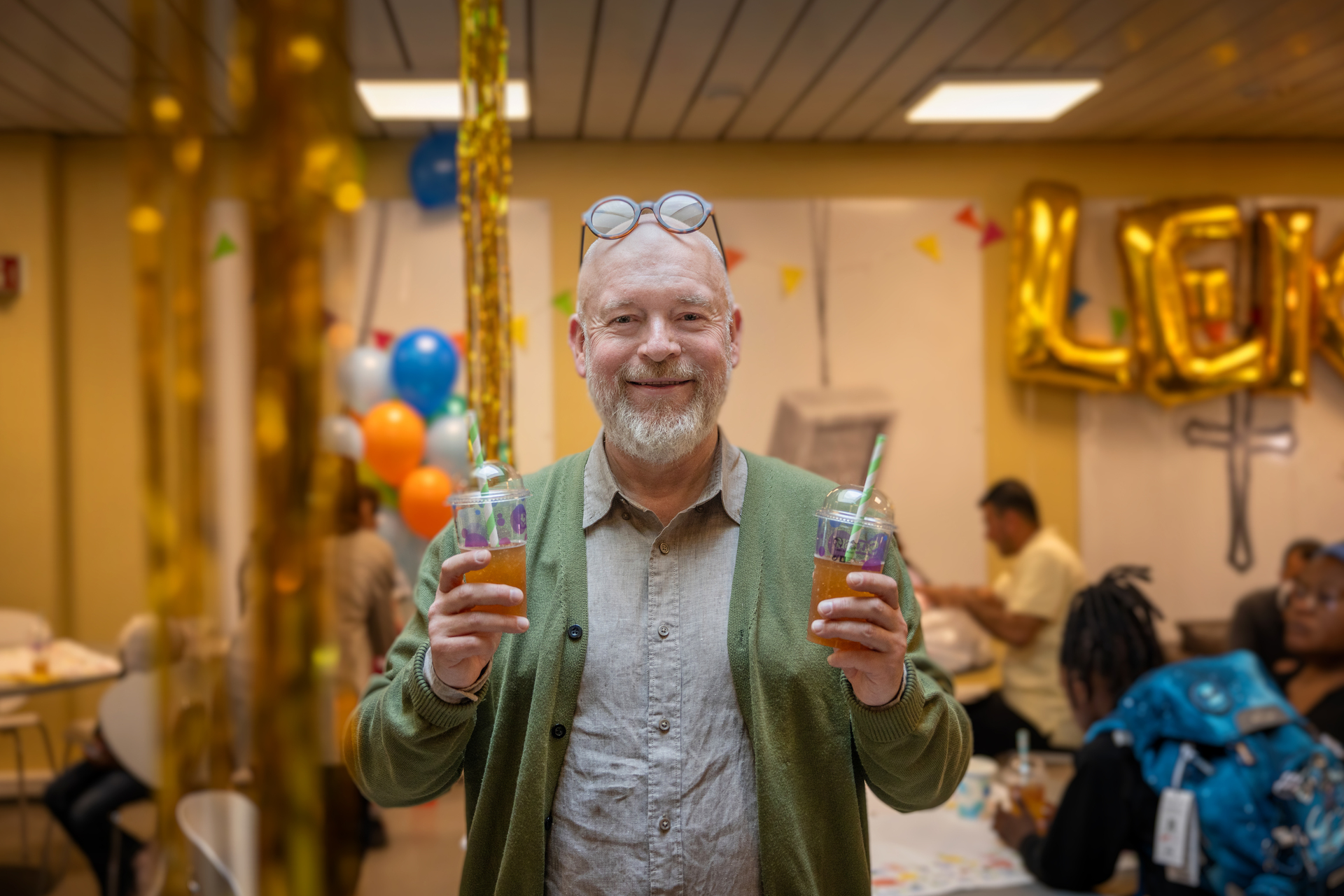 Foto: En mann står og holder et plastkrus med slush i hver hånd. Han er skallet med grått skjegg. Han smiler mot kamera. Bak ham sees ballonger, gullfarget pynt og en gutt med ny skolesekk sees uskarpt i bakgrunnen.