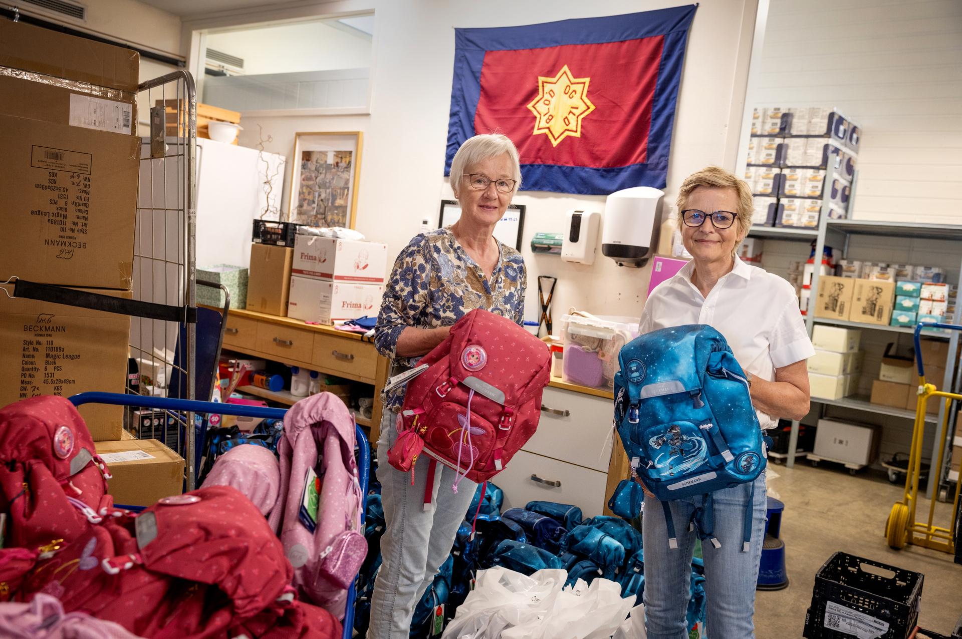 Foto: To kvinner med kort hår og briller står på et lager og holder opp hver sin skolesekk, en rosa og en blå. Bak dem henger et stort Frelsesarme-flagg, og på den ene siden står en tralle med mange flere sekker på.