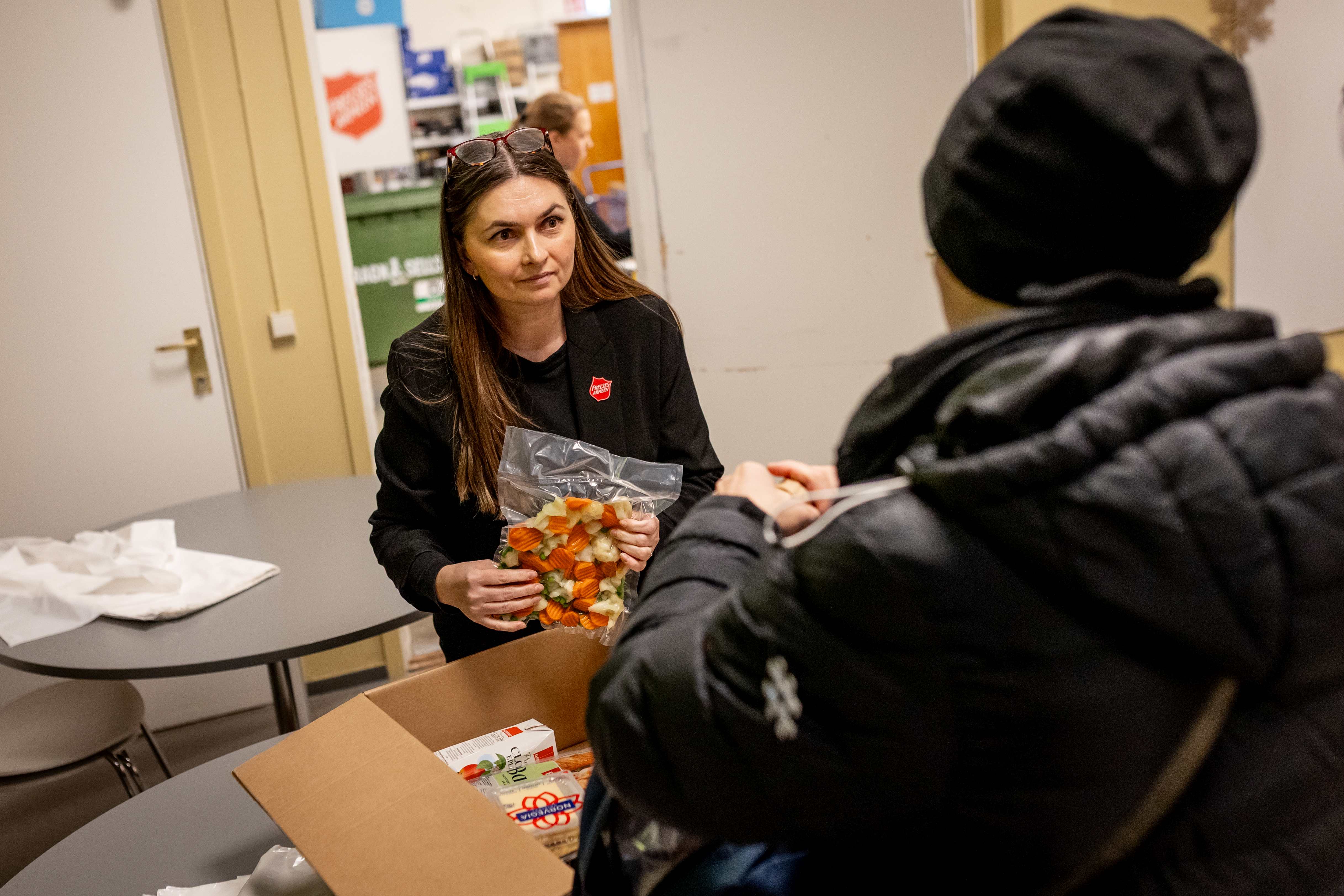 Foto: Dame fra Frelsesarmeen i samtale med en gjest (med ryggen til) inne på slumstasjonen. Mellom seg har de en eske mat