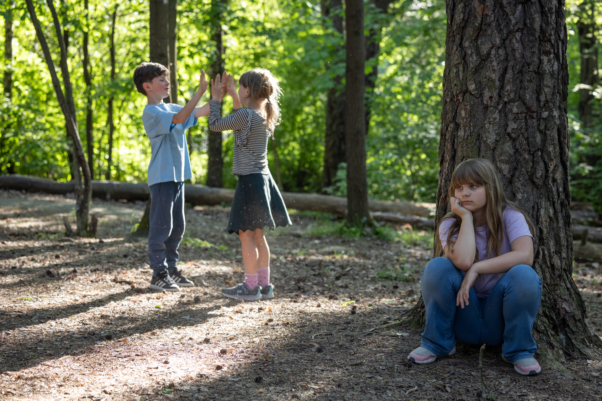 Foto: Tre barn leker i skogen, men ei av jentene er utelatt fra leken og sitter med ryggen mot en trestamme.