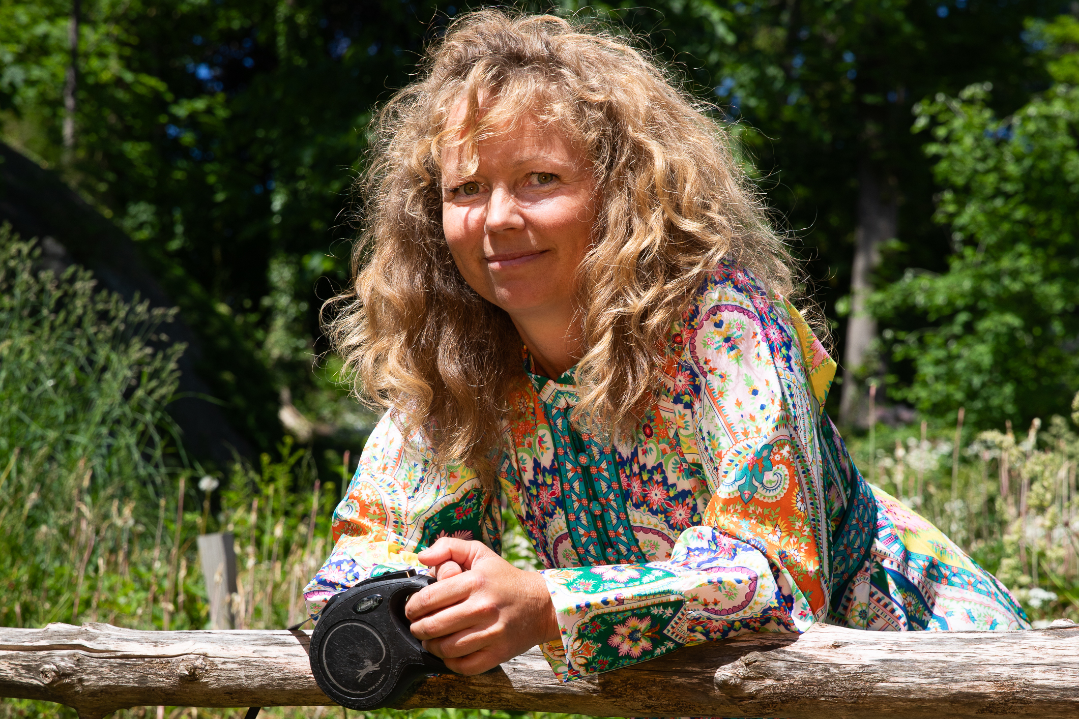 Kvinne med lys krøller og blomstrete sommerkjole lener seg på et gjerde.