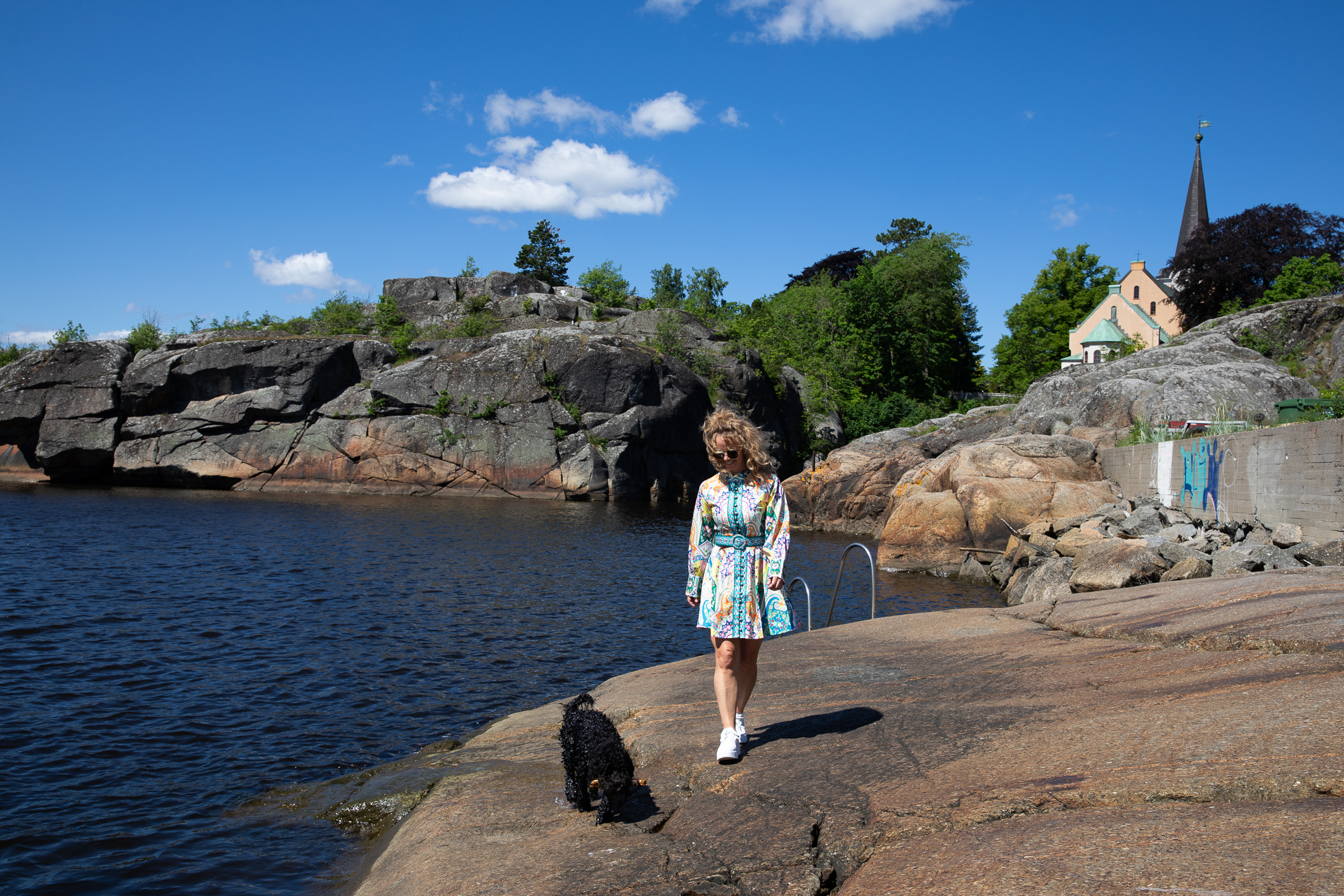 Kvinne med lys krøller og blomstrete sommerkjole på et svaberg sammen med en sort hund. Blå himmel og en kirke i bakgrunnen.