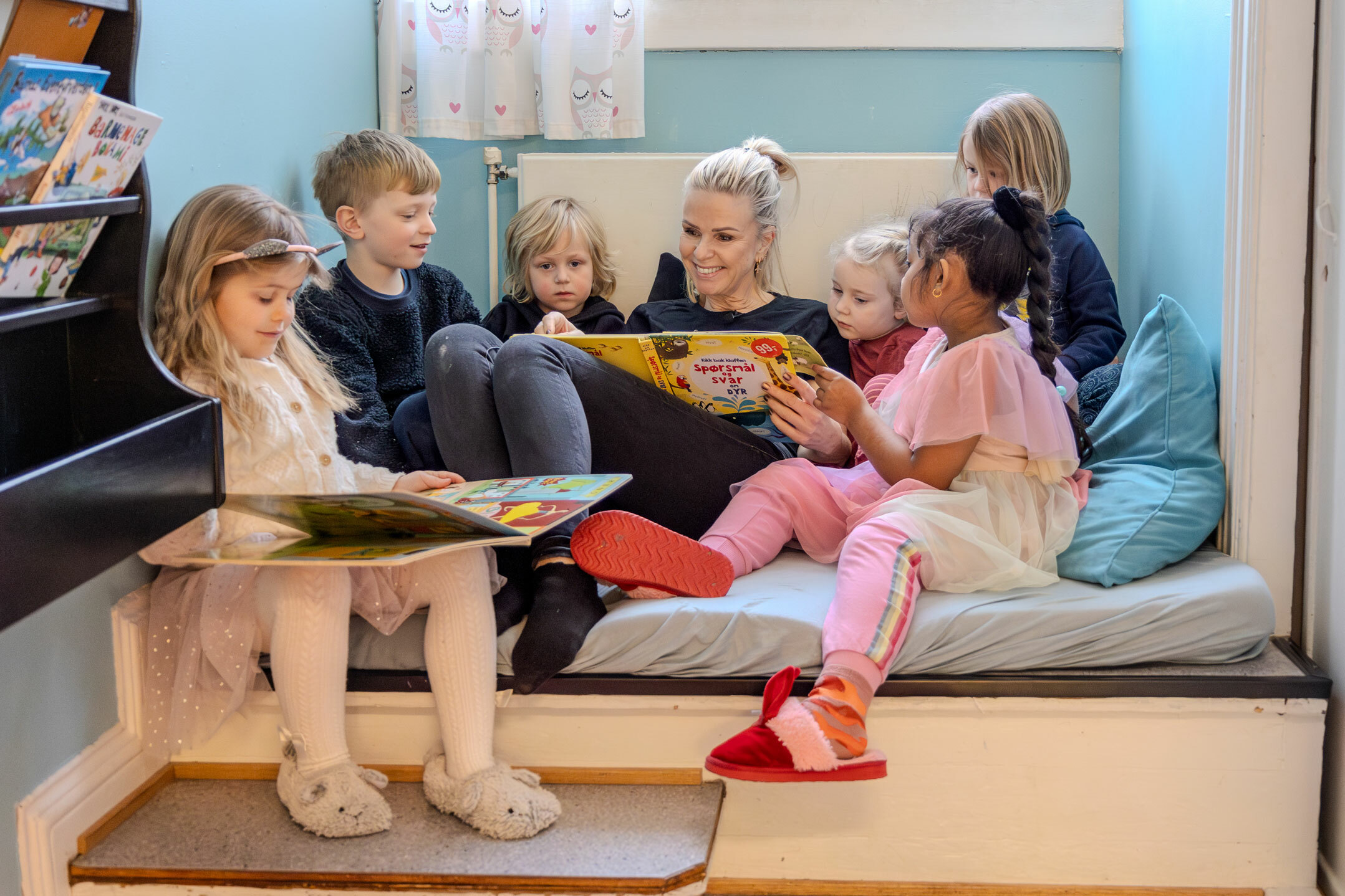 Foto: Voksen barnehagetante sitter i en kosekrok med seks barn – tre jenter og tre gutter. Hun leser i en bok for dem.