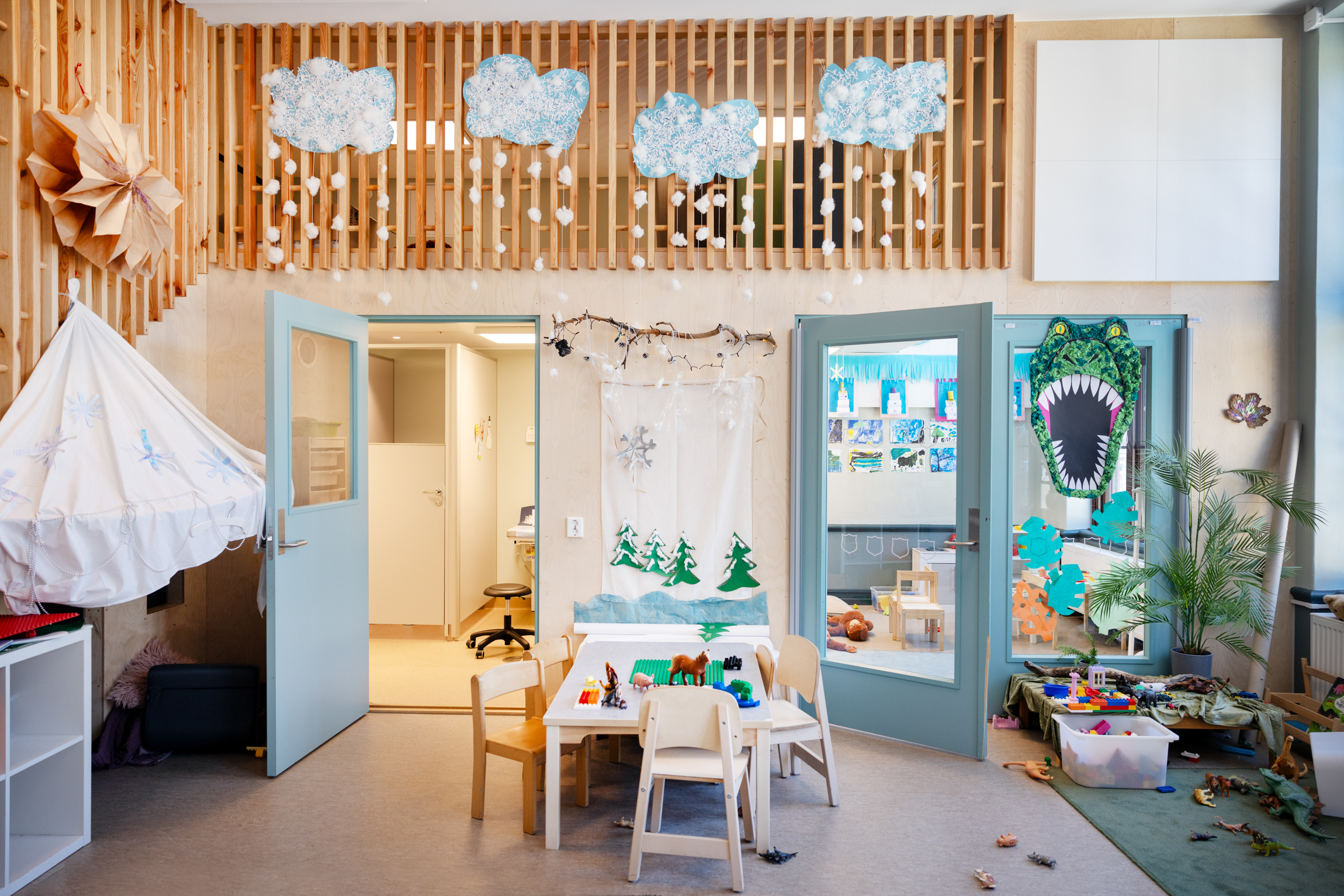 Barnehageavdeling med duse farger på dører, lite trebord med stoler og en masse leker. På et av vinduene inn til neste rom henger et dinosaurhode barna har laget sammen ved å rulle sammen små "kuler" i silkepapir med ulike grønnfarger..