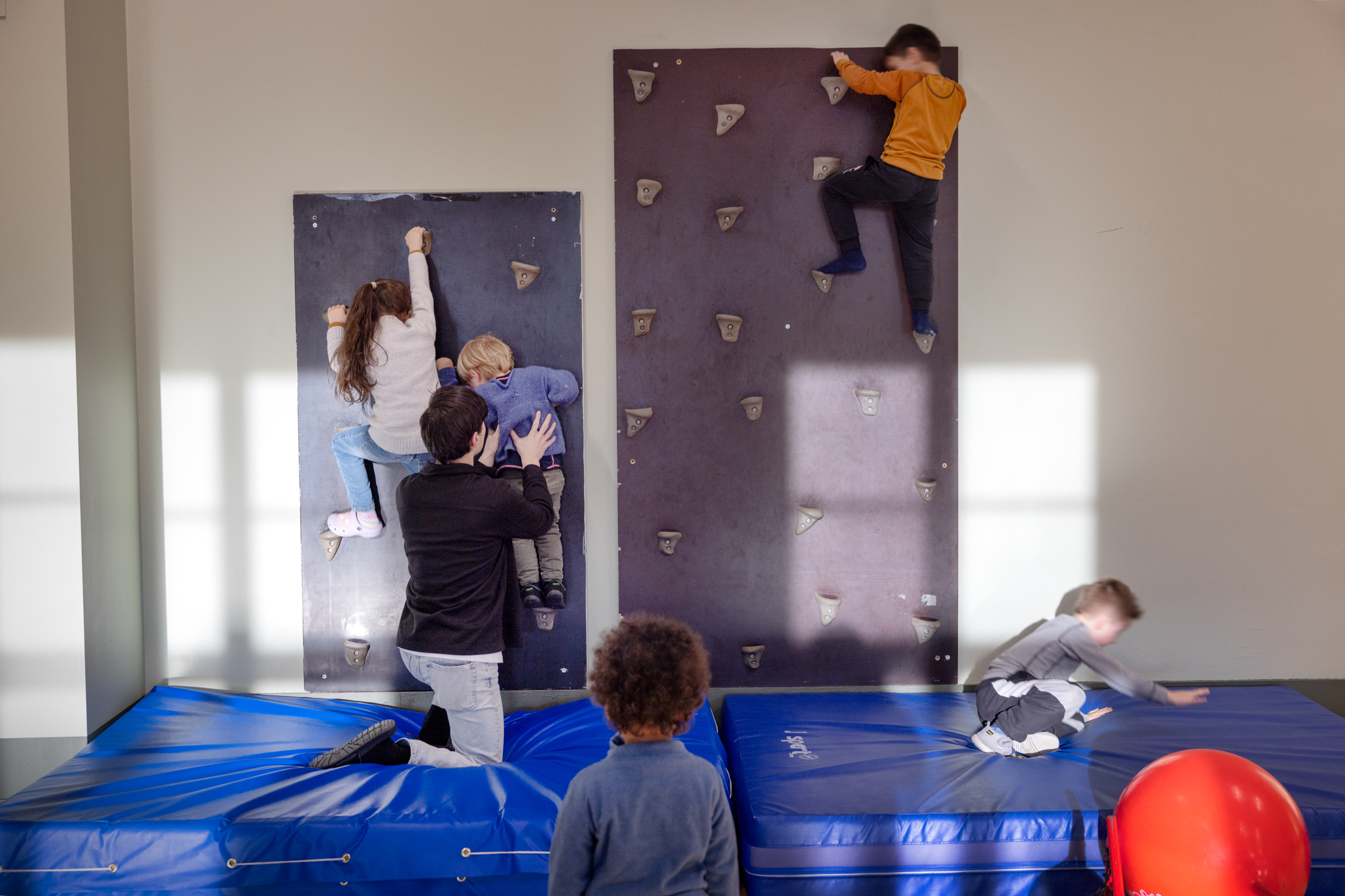 Tre barn som beveger seg oppover i en klatrevegg der den yngste får hjelp av en voksen. To andre barn leker seg på den tjukke blå matta like under.