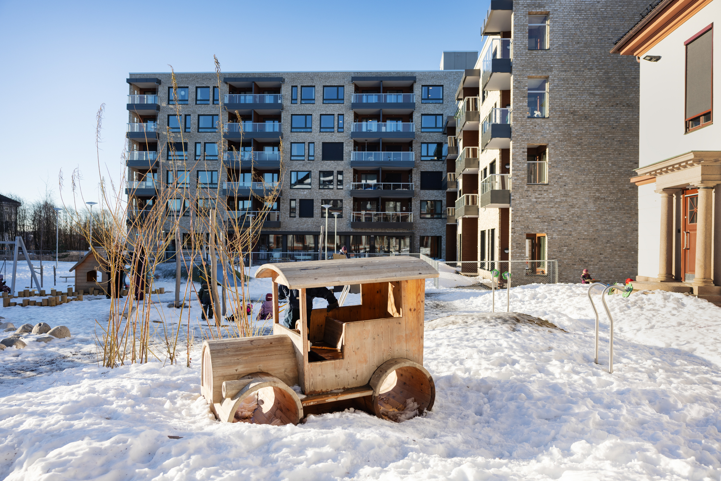Lekeområde med en stor bil i tre som barna kan sette seg inn i. Bakenfor er det mange leiligheter i etasjene oppover.