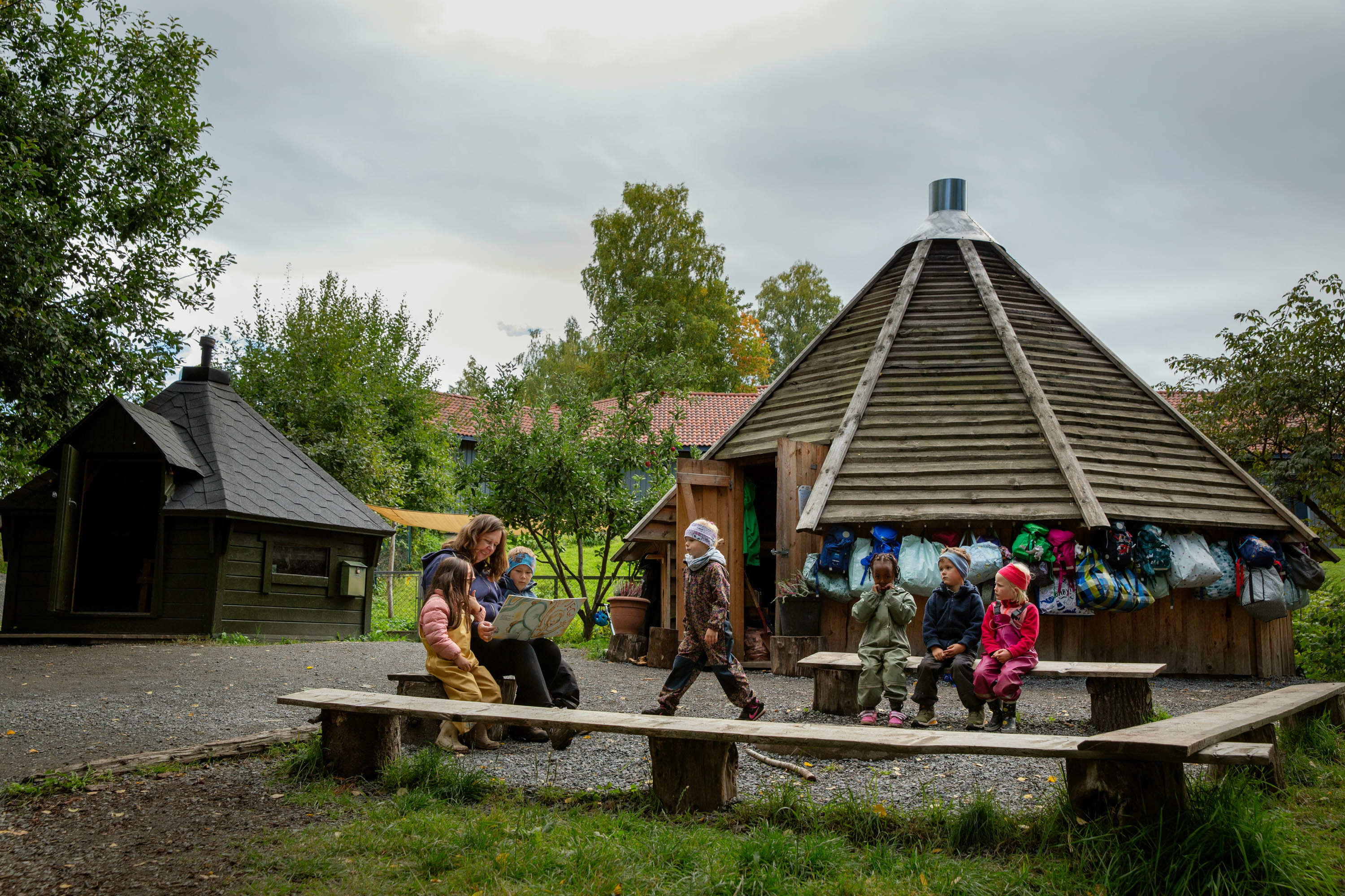 Foto: Barn løper i utetøy foran en åttekantet tømmerkoie og en liten lekehytte ved siden av. Foran den største hytta er det benker i tre som står i en firkant.