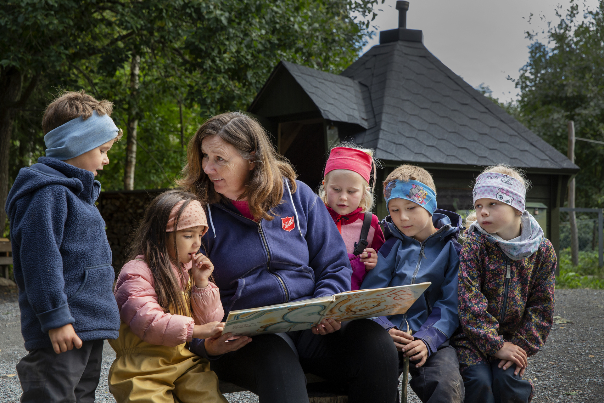Foto: Barnehagetante leser i en bok for fem barn. De er ute og sitter på samme benk.