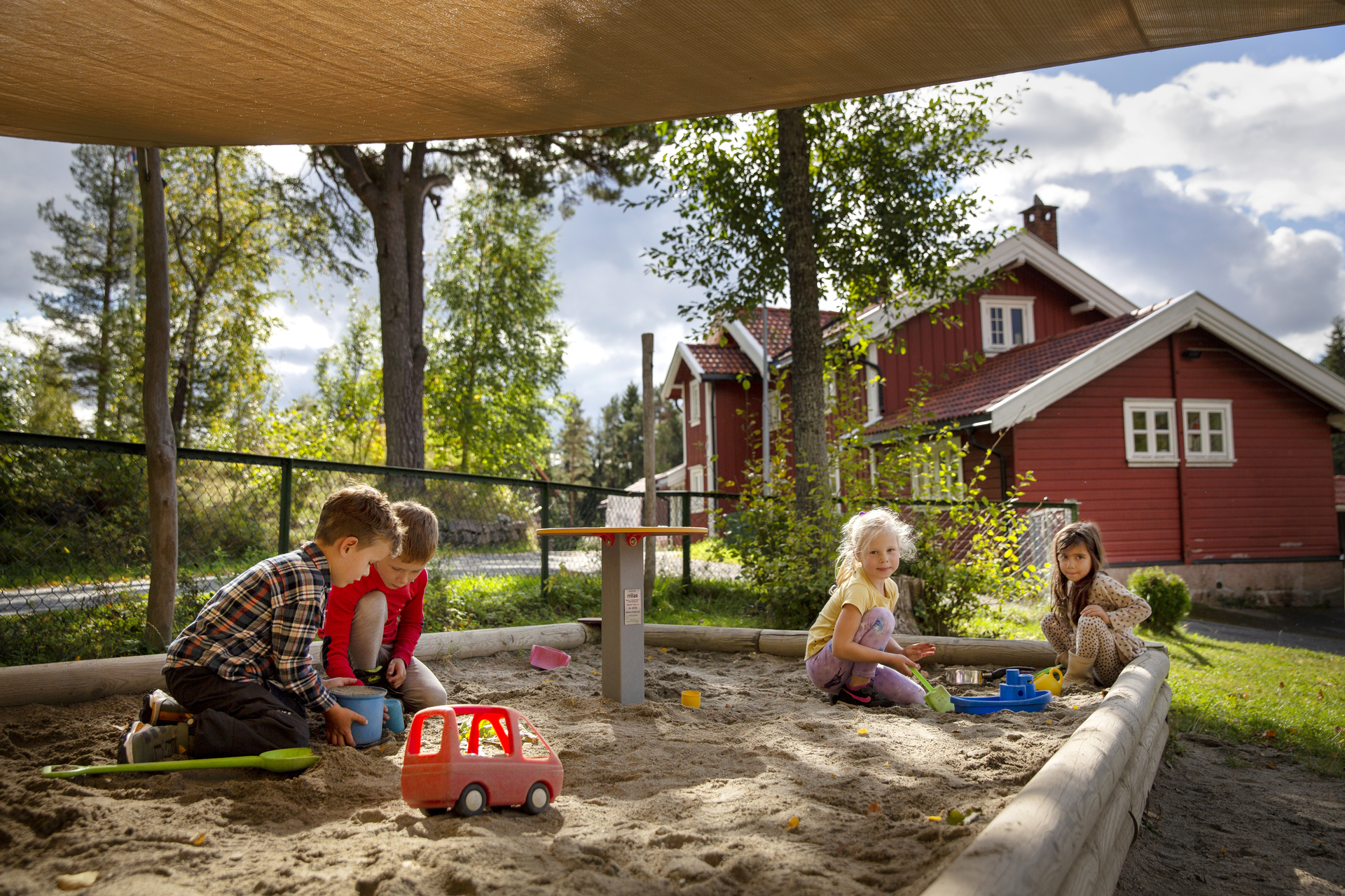 Foto: Fire barn leker i en sandkasse med en seilduk over. I bakgrunnen står et stort, gammelt rødt hus med hvite karmer. Rundt er det grønne områder med gress og trær.