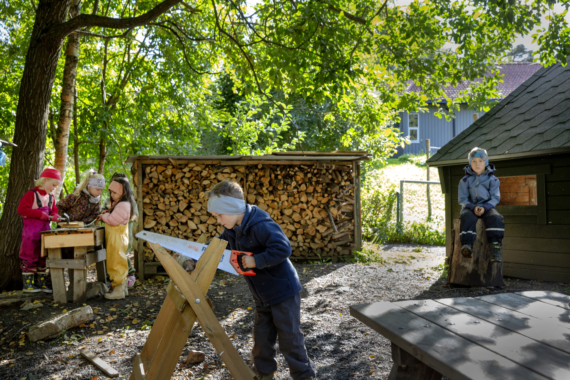 Foto: To bilder etter hverandre, der det første viser tre jenter som spikrer på et bord og dena ndre bildet viser en gutt som sager. I bakgrunnen er det stablet tre i et lite hus med tak over.