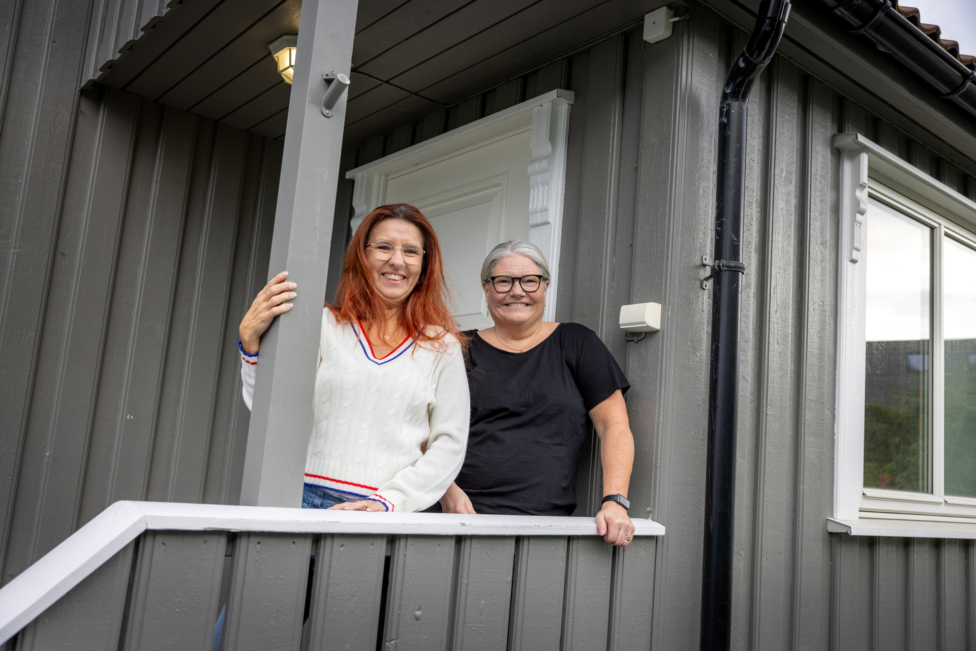 Foto: To voksne kvinner står utenfor inngangspartiet med rekkverk opp til et grått hus med hvit dør og hvite vinduskarmer.