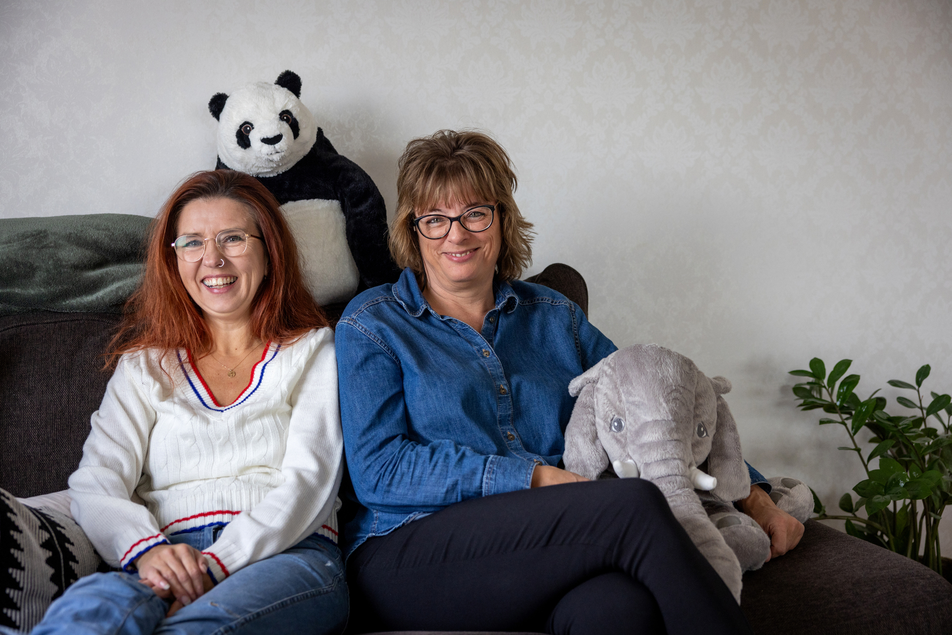 Foto: To voksne kvinner sitter i en sofa sammen ned et par kosedyr. Begge har briller og begge smiler til fotografen.