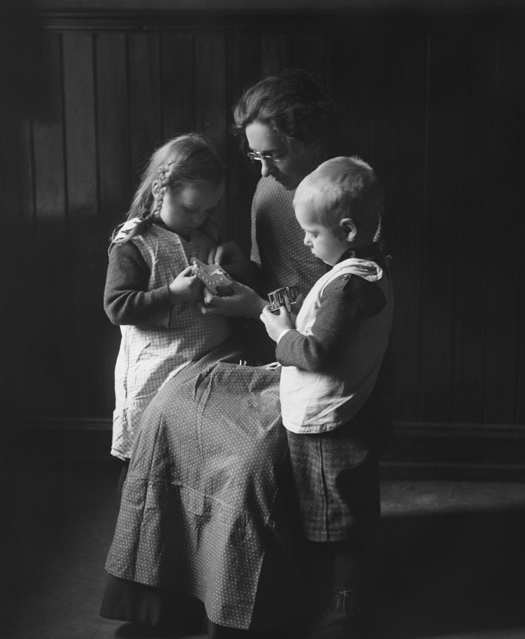 Foto: I svart hvitt fra "gamledager" der en kvinne i svart kjole med hvitt forkle og oppsatt hår sitter på en stolt. Rundt hennes står det to barn.
