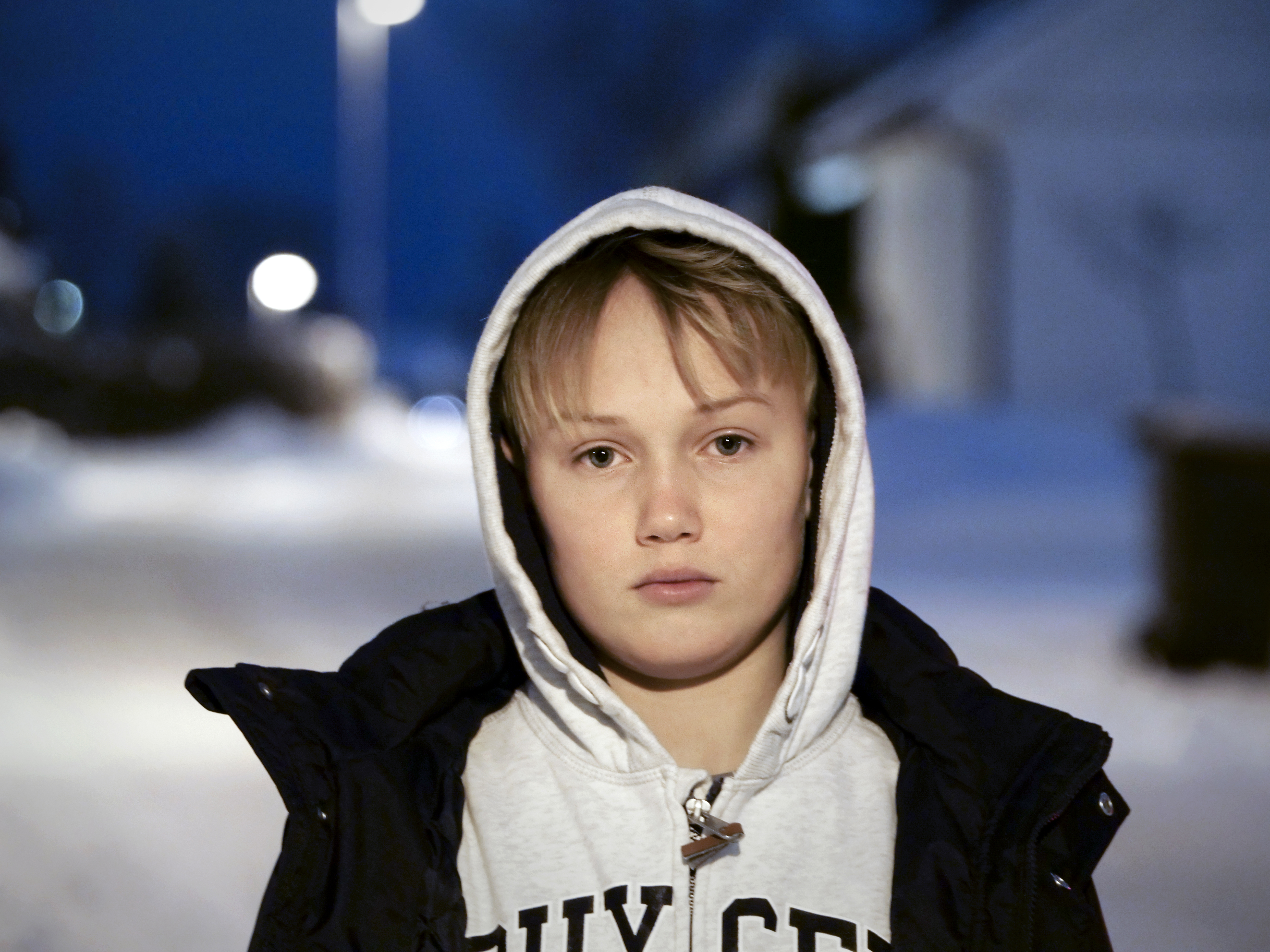 Lyslugget gutt står utendørs og ser rett inn i kameraet. Har på seg en hettejakke og en ytterjakke.