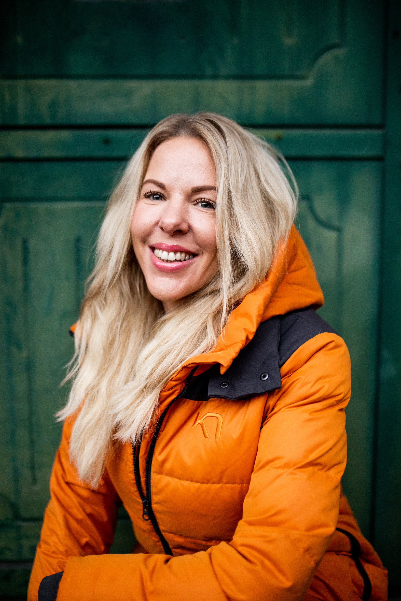 Gatefotballspiller og -trener Cathrine Johansen står foran en grønn dør. Hun har blondt, langt hår og smiler mens hun ser inn kameraet. Hun har på seg en knall oransje dunjakke.