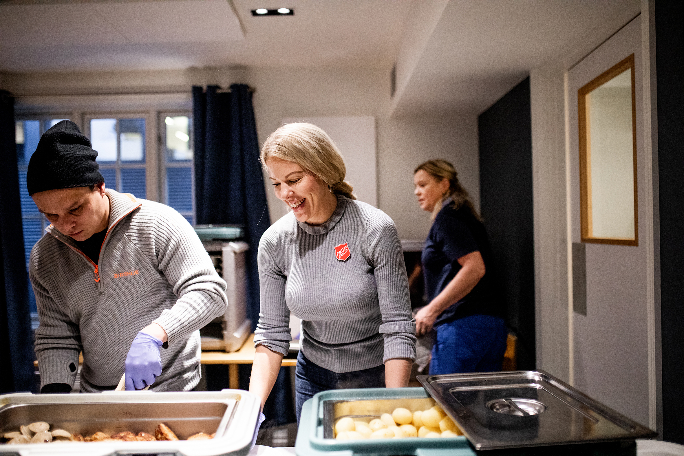 Cathrine står sammen med to andre og forbereder matservering på Stedet Sandvika. Bakker med poteter og annen mat man ikke kan skimte, ses så vidt i forgunnen på bildet. Som miljøarbeider på Stedet Sandvika sørger Cathrine (t.h.) for at alle trives. (Foto: Kristianne Marøy)
