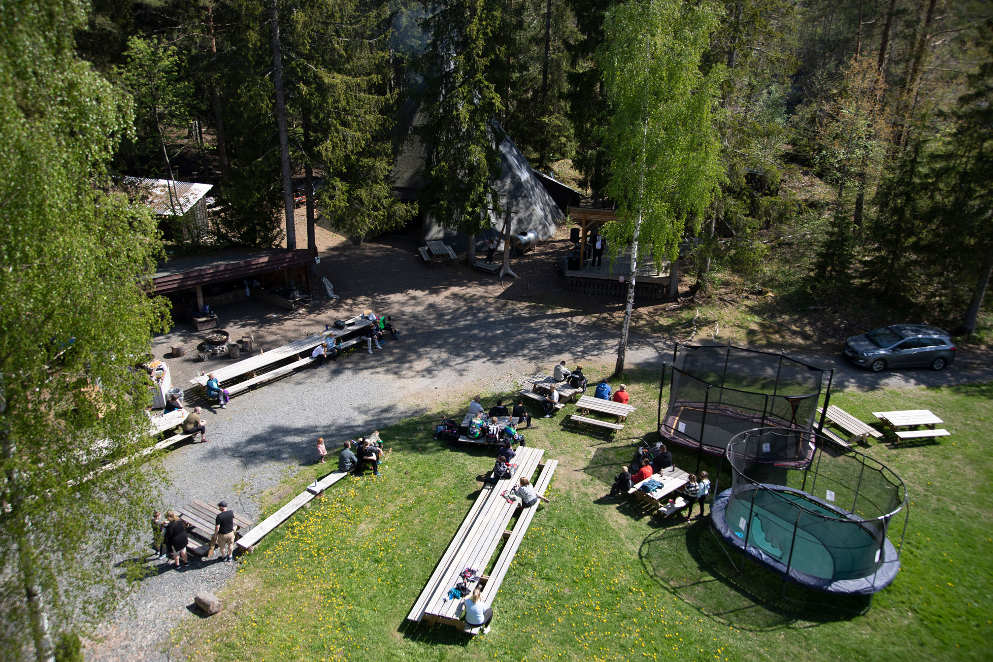 Foto: Oversiktbilde av en villmarkscamp-leir med trampoliner, benker og bord. Ungdommer sitter ved noen av bordene.