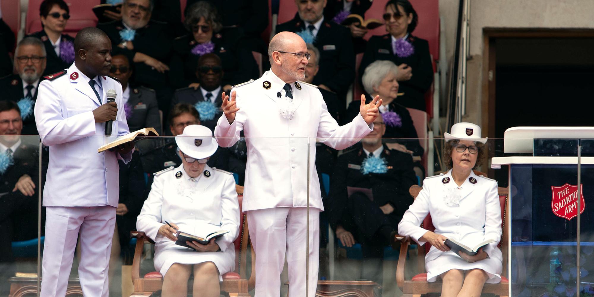 4 bilder: 1. Generalen i hvit uniform står på talerstolen på et utendørs stadion. 2. Ung afrikansk kvinnelig soldat danser foran mange andre unge soldater. 3. Hornorkester i røde jakker marsjer inn på stadion, Kenyas og Frelsesarmeens flagg bæres foran. Leder og nestleder med koner, alle i hvite uniformer marsjerere inn på stadion