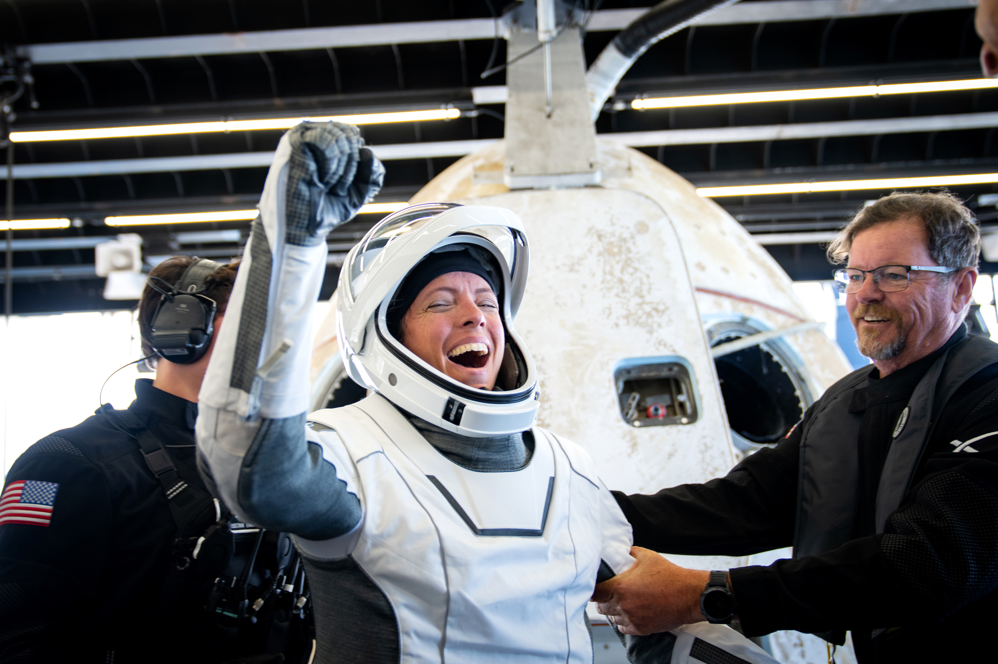 Foto: En kvinne i astronautdrakt holder en arm med knyttet neve i været mens hun smiler med lukkede øyne. En mann i svarte klær holder henne i armen og smiler mot henne, en annen mann med amerikansk flagg på armen skimtes i bakgrunnen, foran deler av romfartøyet.