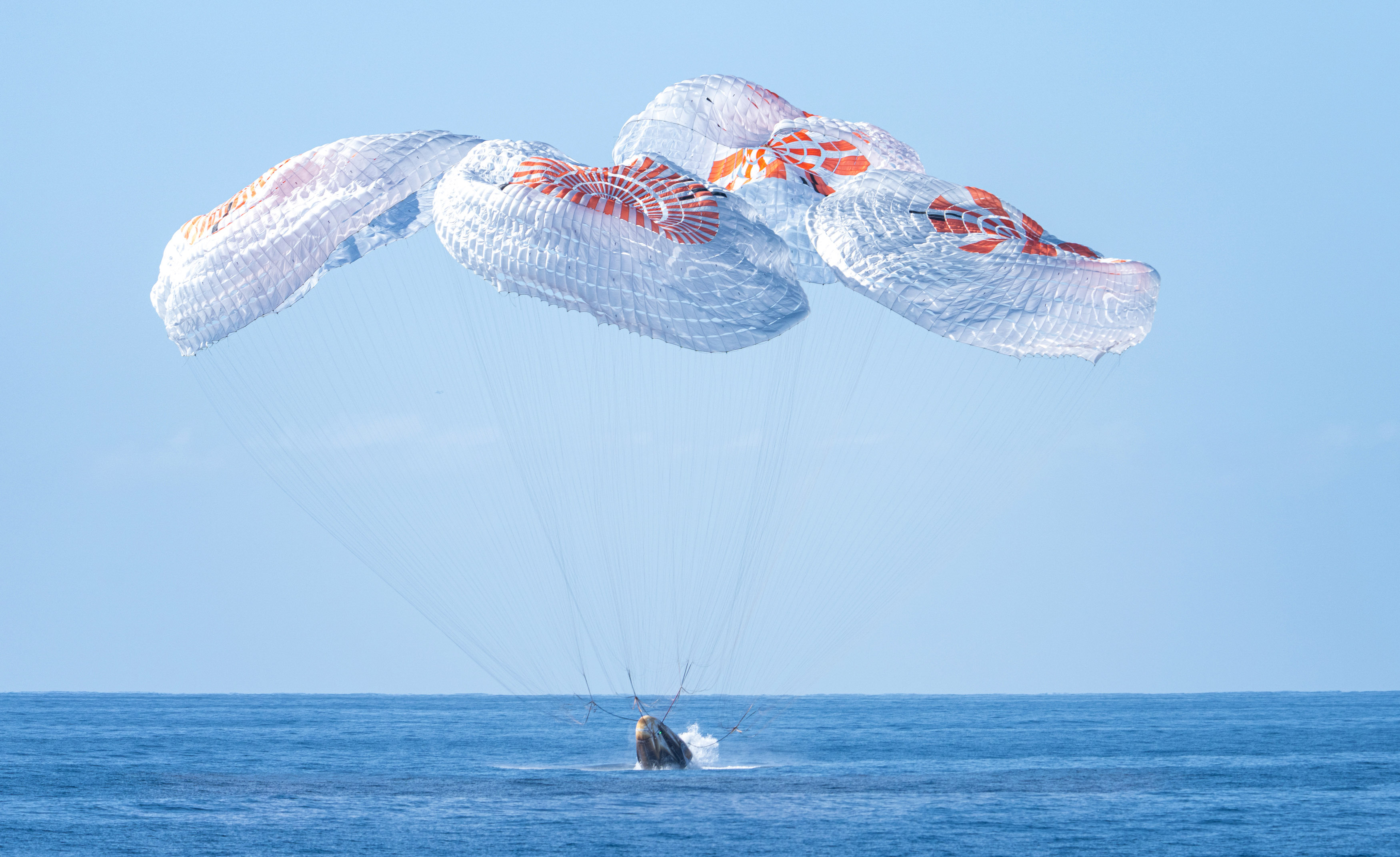 Foto: Romfartøyet treffer vannoverflaten, med flere store fallskjermer over seg. Bare vann og blå himmel sees ellers i bildet.