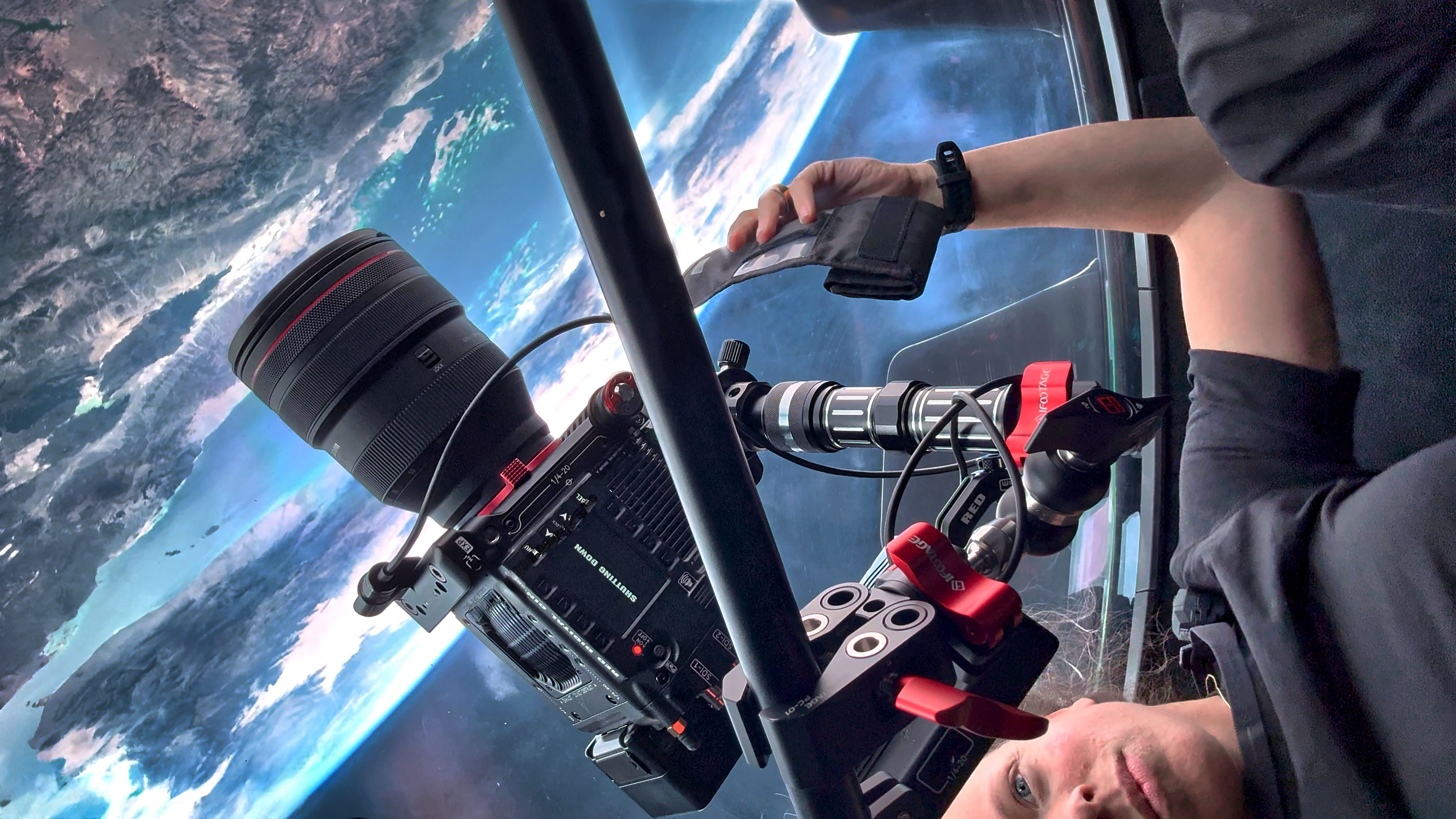 Foto: En kvinne holder et stort kamera, hun sees nederst i høyre hjørne på bildet. Utenfor et vindu sees deler av jorden, sett fra verdensrommet.