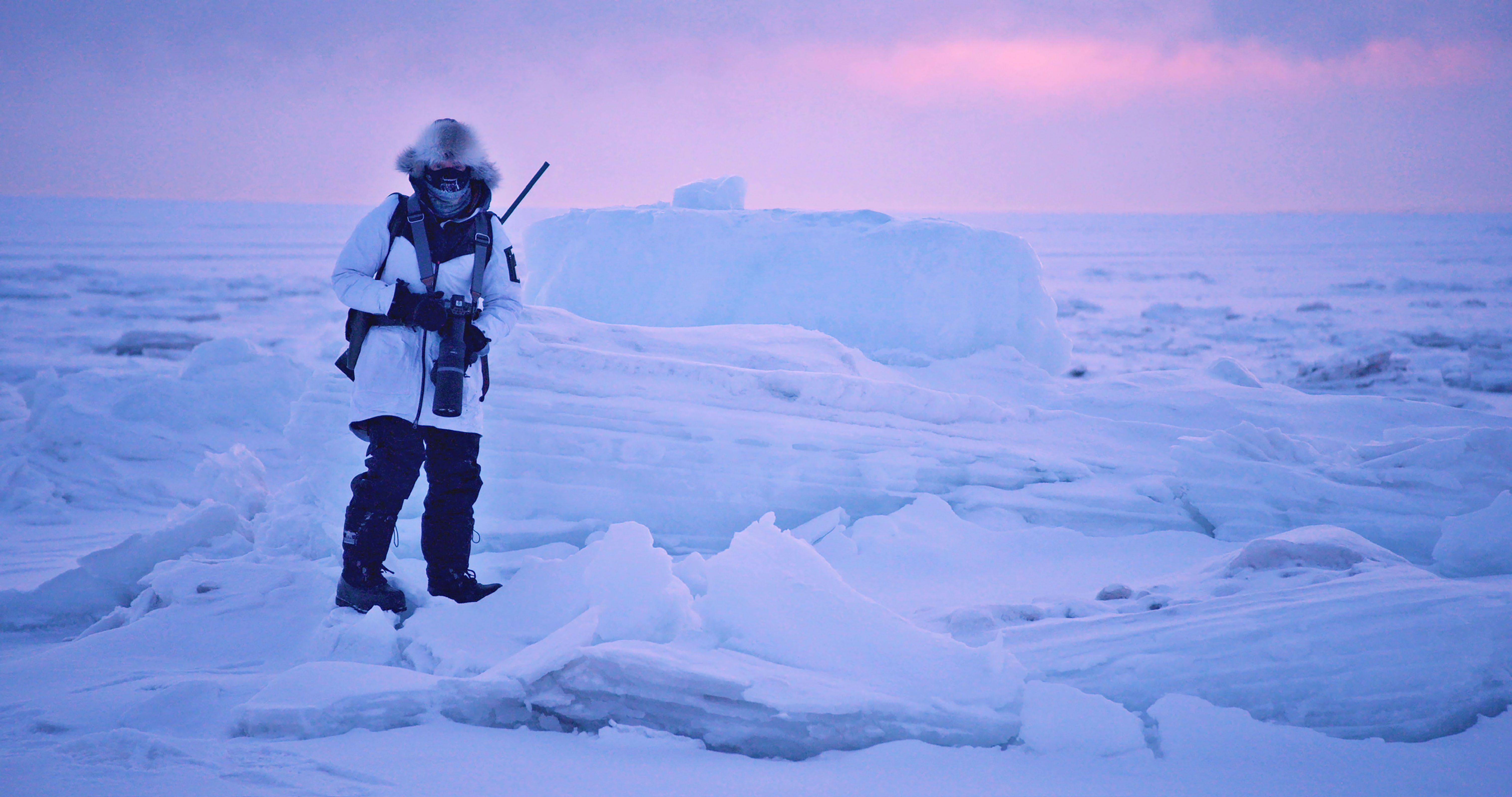 Foto: En kvinne står blant store isblokker og snø, i vinterlys med rosa himmel i bakgrunnen. Hun har på seg hvit jakke med hette, og ansiktet er dekket med et skjerf.