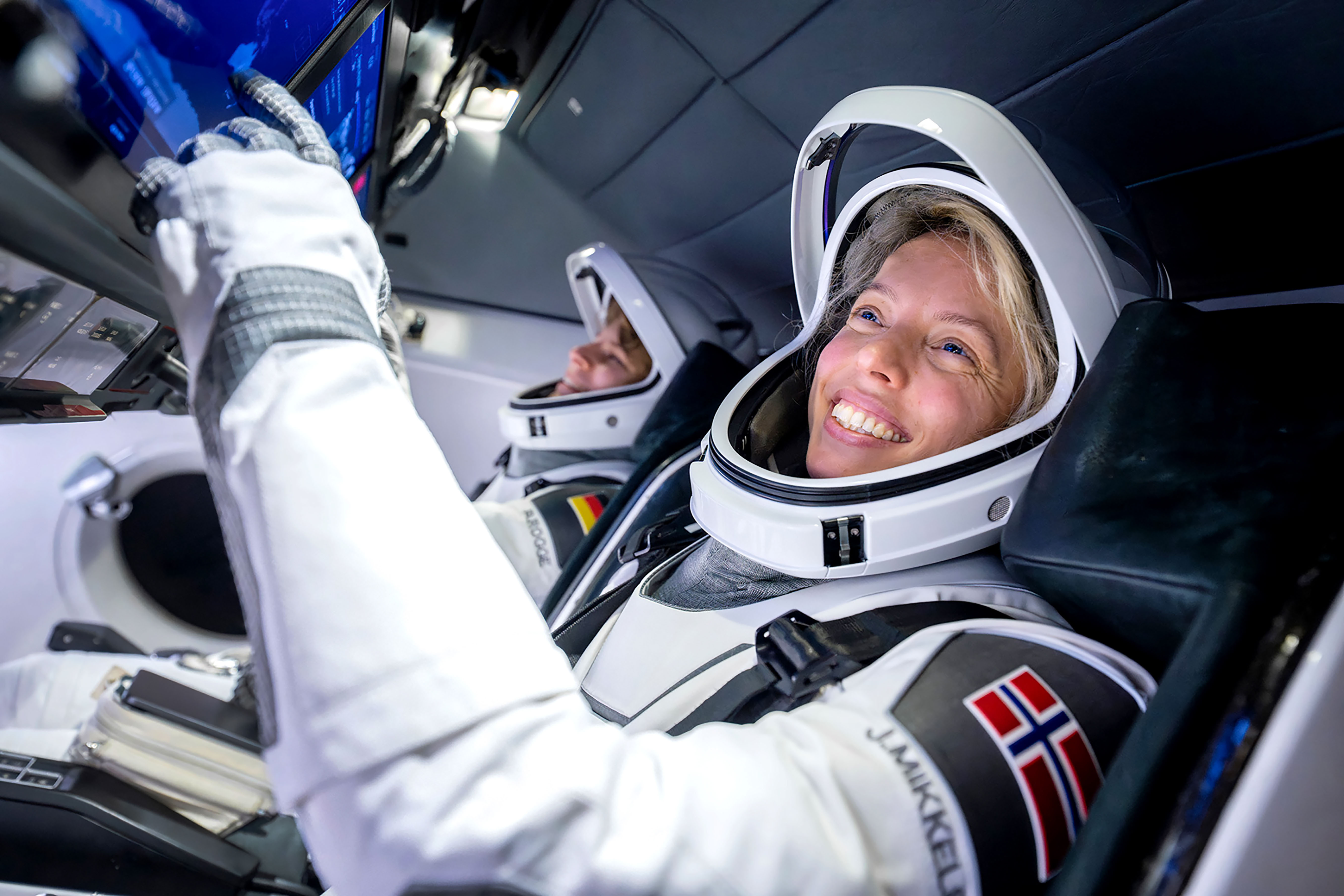 Foto: Bildet er tatt fra siden og viser to personer i astronautdrakt inne i et fartøy. Jannicke sitter nærmest og smiler mens hun ser på noe foran seg. Den andre personen er uskarp i bakgrunnen.