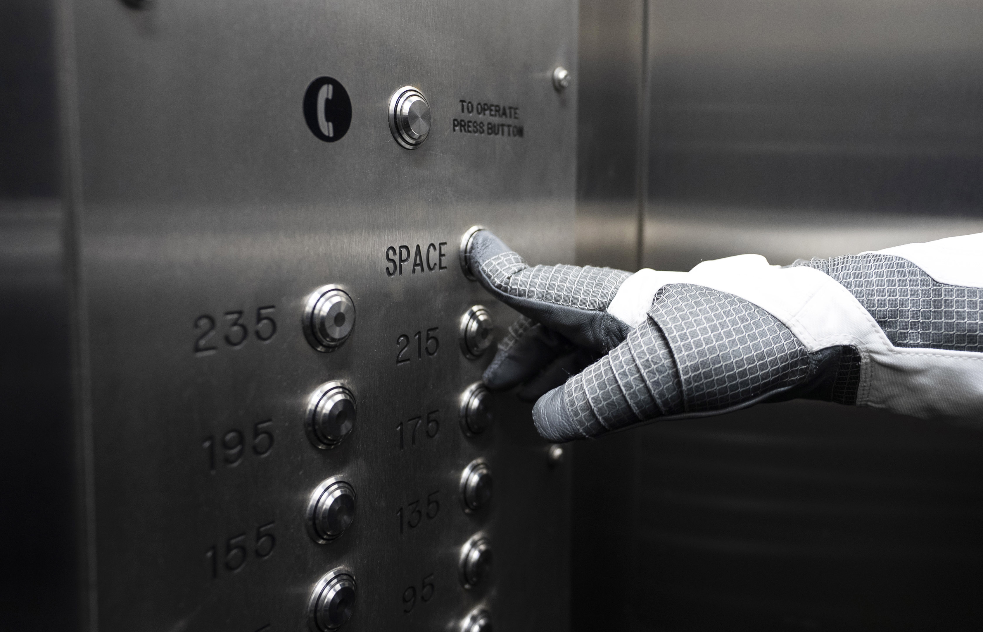 Foto: En hånd med hvit hanske trykker på den øverste knappen på en metallvegg i det som ser ut som en heis. Over knappen står det "space".