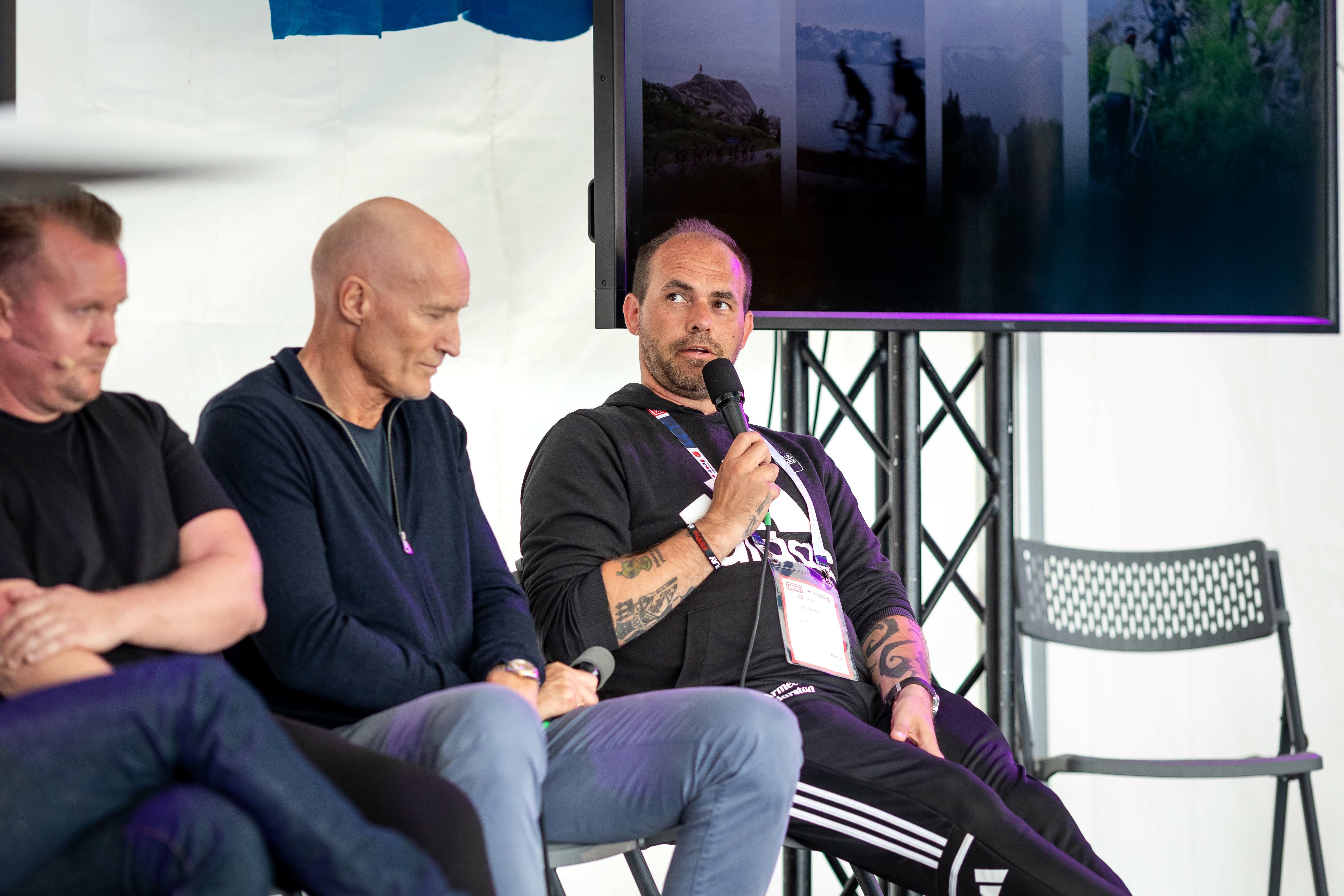 Foto: Helge Dahlberg i svarte klær sitter på en scene og holder en mikrofon. Ved siden av ham sitter to middelaldrende menn, også i svarte klær. Begge ser ned og ser ut til å lytte.