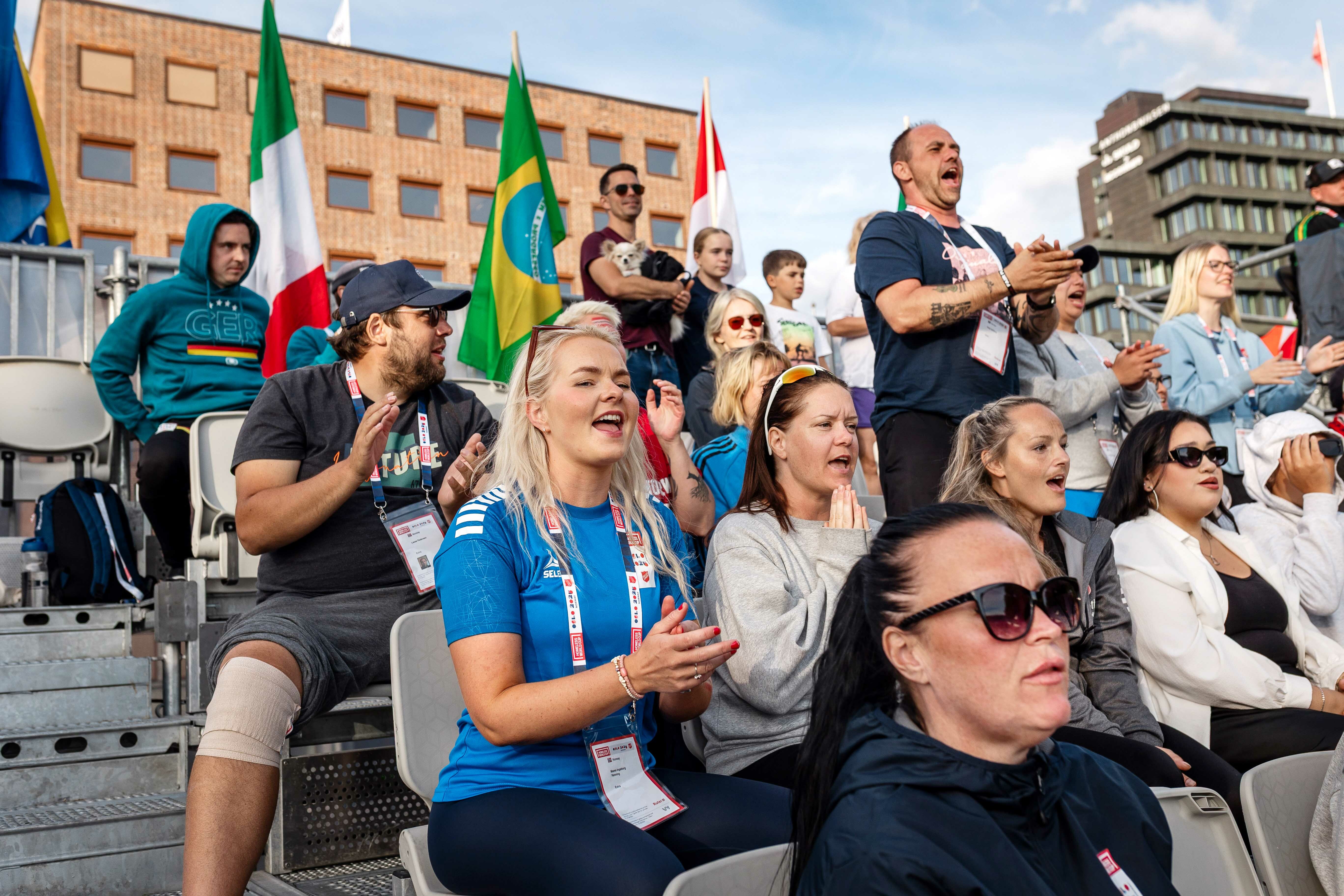 Foto: En gruppe kvinner og menn sitter på en tribune. Flere ser ut som de roper og klapper. De fleste sitter, Helge Dahlberg har reist seg opp og ser ut som han roper mot banen.