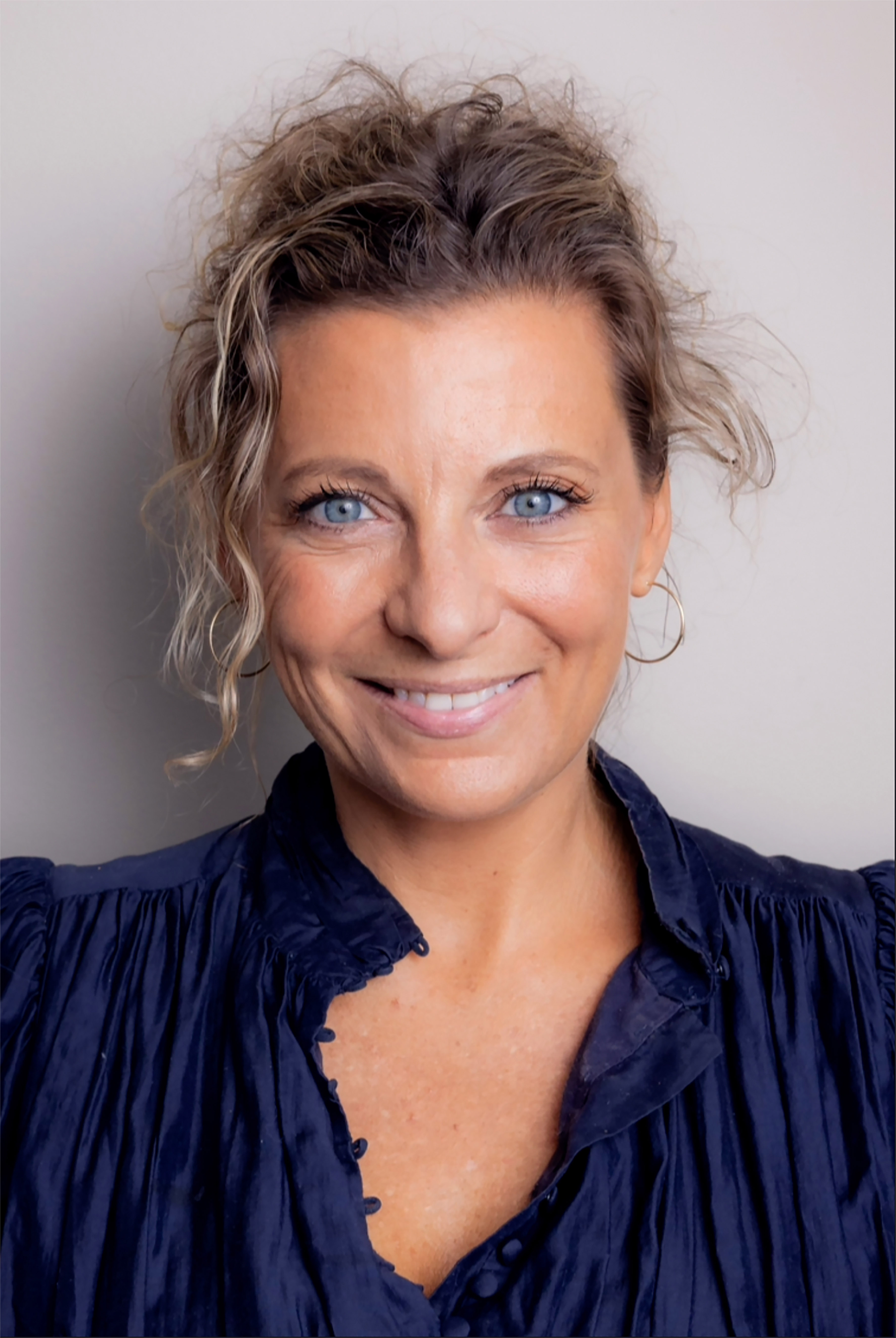 Foto: Portrett av en kvinne som smiler mot kamera, hun har brunt, bølgete hår løst oppsatt, og mørk ble skjorte.